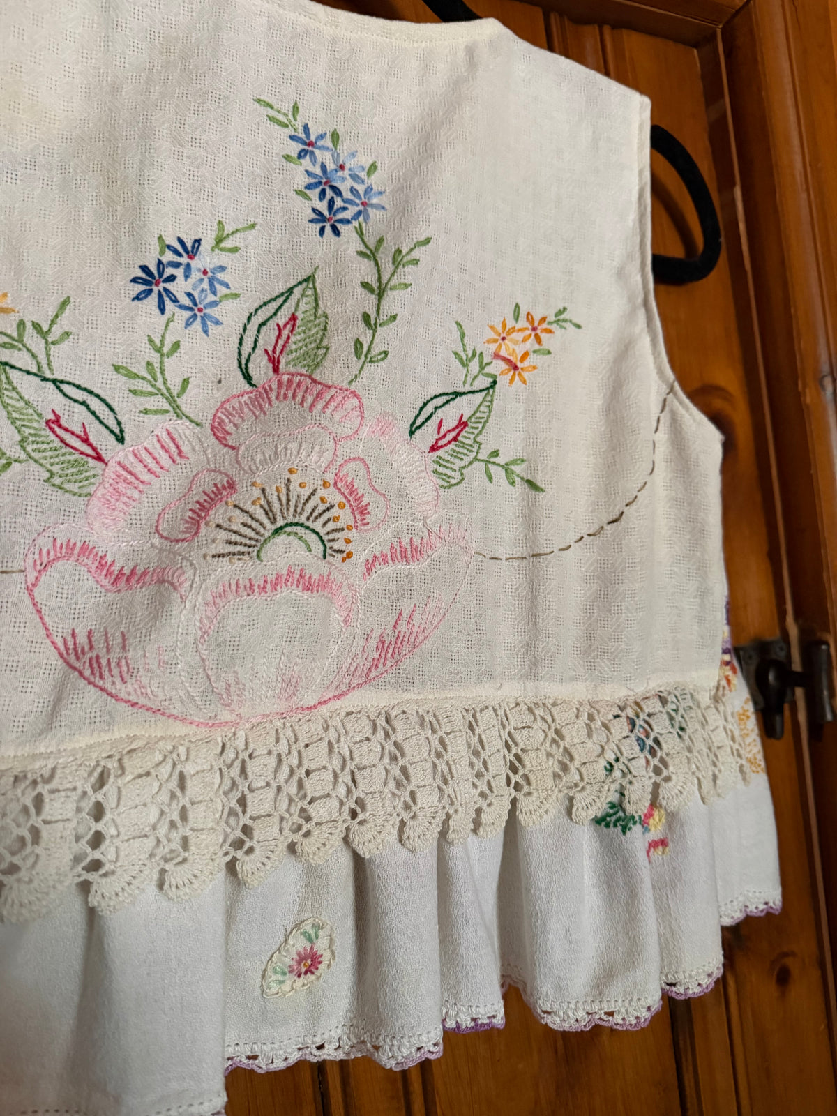 An off-white linen vest with colorful floral embroidery and a lace hem, hanging on a black hanger against a wooden door background.