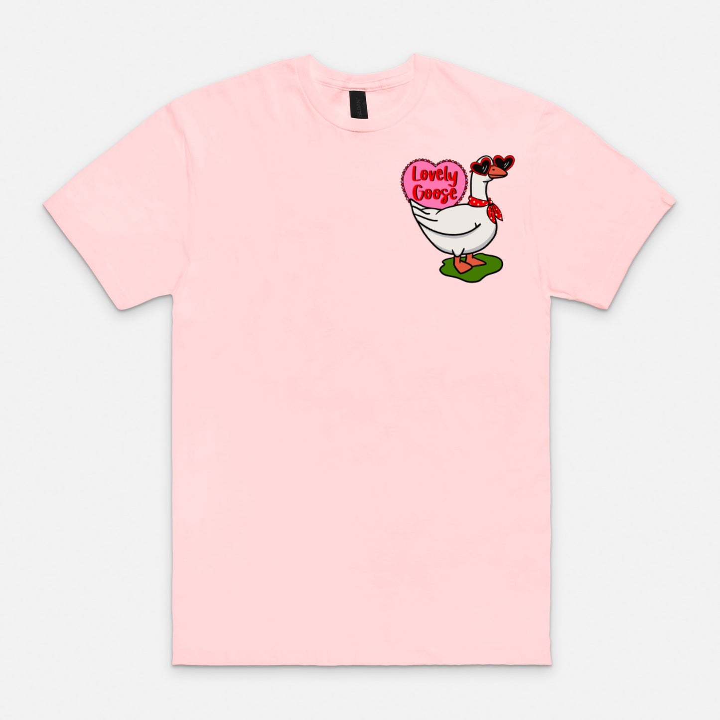 Porch goose pink t-shirt featuring porch goose in heart shaped sunglasses and red cloth tied around the neck with green patch under its feet. A pink scalloped heart partially covered by goose with text inside the heart that reads, “Lovely Goose”. Design. Is a smaller chest piece. Plain white background flat lay.