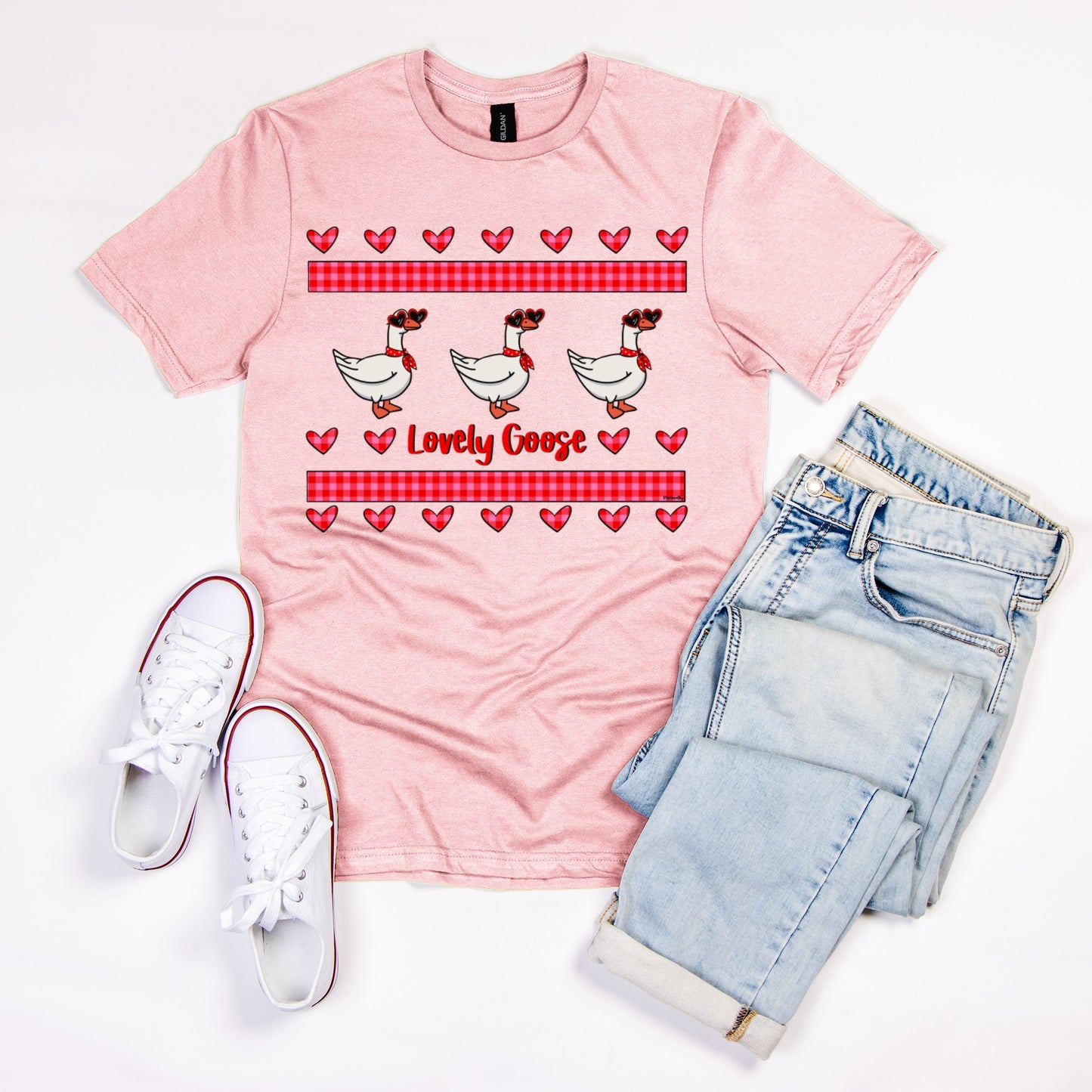 Porch goose pink T shirt featuring three porch geese in heart shaped sunglasses and red cloth tied around the neck with plaid pink and red rectangles above and below and several hearts creating a love design. Text reads, “Lovely Goose”. Flat laid with jeans and white sneakers.