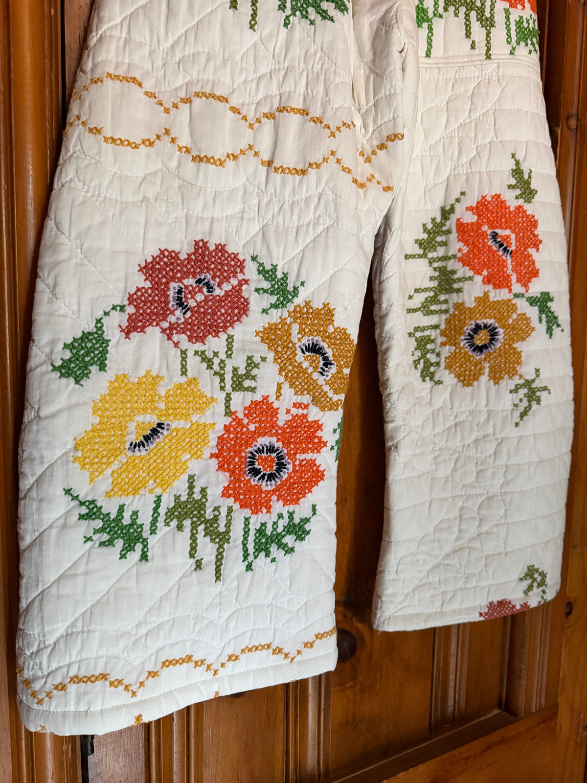 A quilted cream-colored apron or bib with colorful embroidered poppy flowers in red, orange, and yellow, hanging on a wooden door.