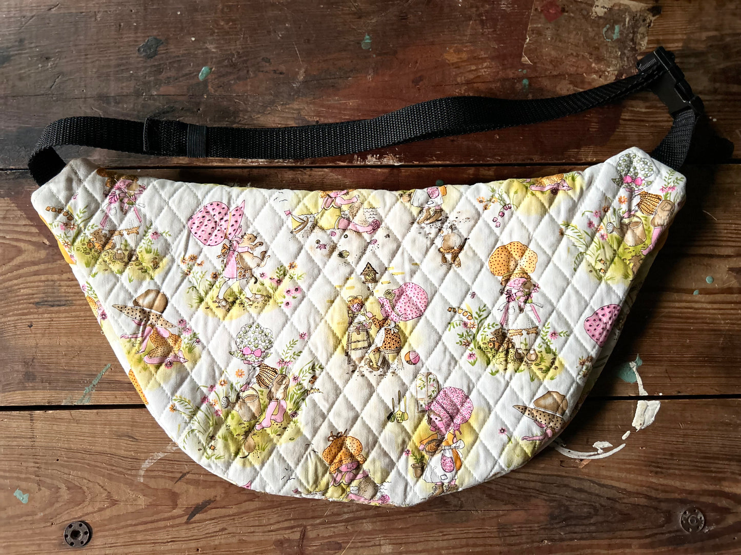 Multicolored Quilted Cross body bag in two tone vintage quilt and quilted fabric. Bottom plaid portion is predominantly white, yellow with touches of pink. The bottom quilt portion contains a vintage style girl in oversized hats in various activities such a carrying a large cat, gardening, playing in sand and playing with toys. Backside view constructed with vintage girl quilted fabric also showing buckle style closure with nylon strap. Photographed on a rustic wooden surface.