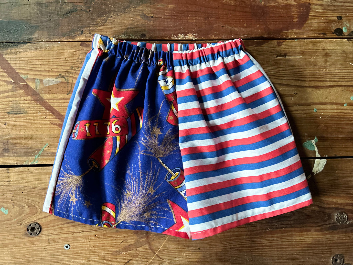 Pair of children’s red, white and blue shorts. Front panels are different with one side featuring lit firework fuses with gold details and 1776 firework. The other front panel features a red, white and blue stripe in a horizontal stripe.