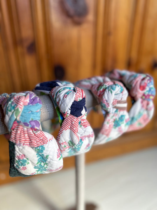 Image is of four knot headbands constructed with a vintage multi color quilt. Primary colors consist of a base color of white with red, greens and blues. The headbands are hanging on a burlap covered necklace holder.
