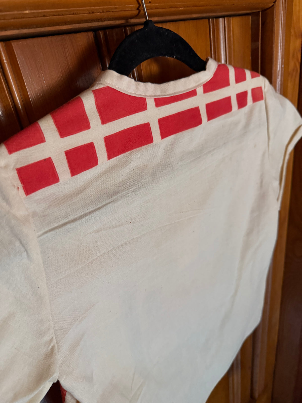 A white short-sleeve shirt with a red grid pattern across the shoulders, hanging on a black hanger against a wooden door.