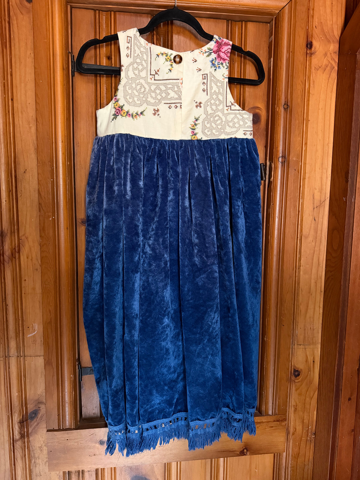 A sleeveless children's dress hanging on a black hanger against a wooden door. The dress has a cream embroidered bodice with floral details and a pleated royal blue velvet skirt with fringe at the hem.