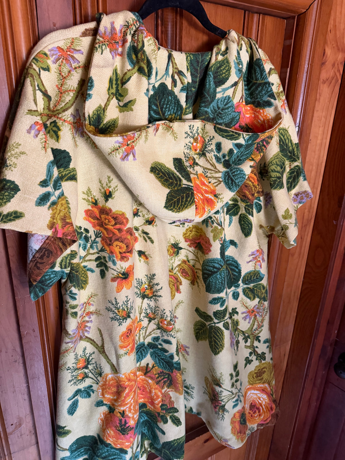 A short-sleeve blouse with a dramatic draped neckline, featuring a vintage floral print on a cream background with orange, red, and green roses and leaves, hanging on a black hanger against a wooden door.