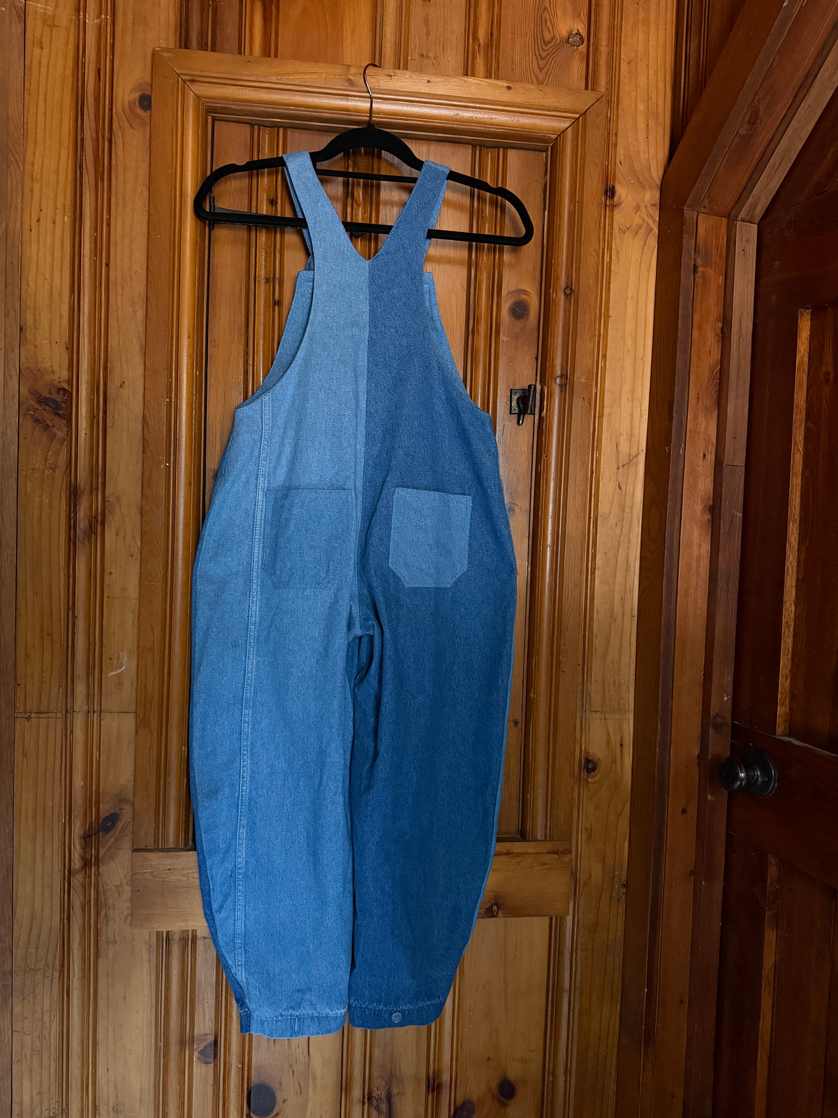 A pair of blue denim bib overalls hanging on a black hanger against a wooden door.