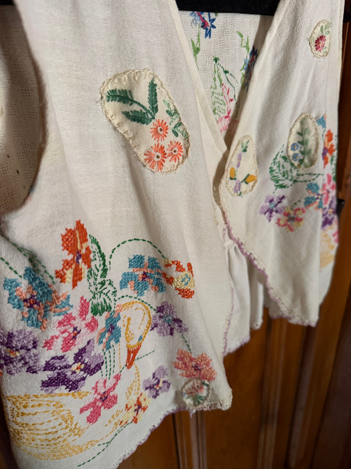 A cream-colored linen vest featuring colorful floral and bird embroidery, hanging against a wooden background.