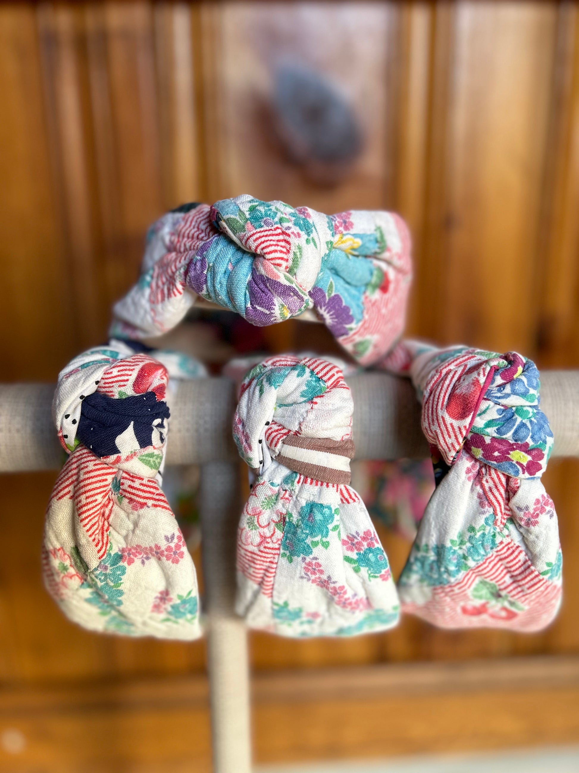 Image is of four knot headbands constructed with a vintage multi color quilt. Primary colors consist of a base color of white with red, greens and blues. The headbands are hanging on a burlap covered necklace holder with one sitting on top of the three headbands showing a forward facing knot for all headbands.