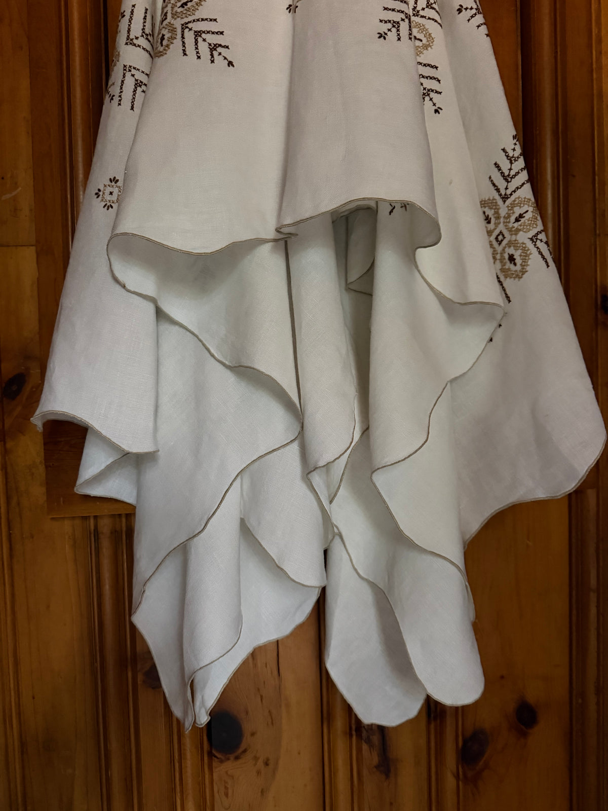 An off-white high-low dress with layered ruffles and delicate brown floral embroidery, hanging against a rustic wooden background.