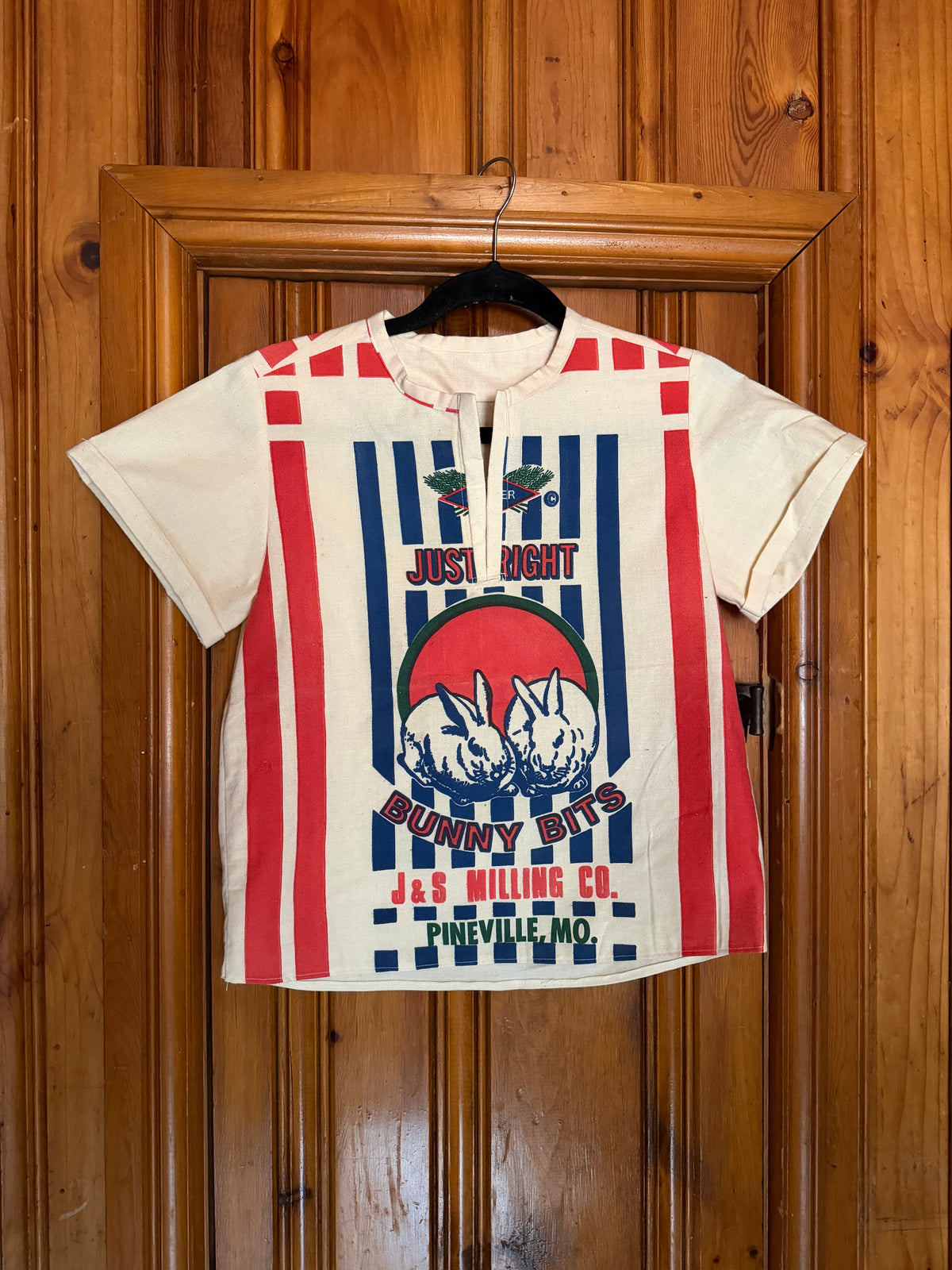 A vintage-style short-sleeve t-shirt with a patriotic red, white, and blue bunny graphic, hanging on a black hanger against a wooden door.
