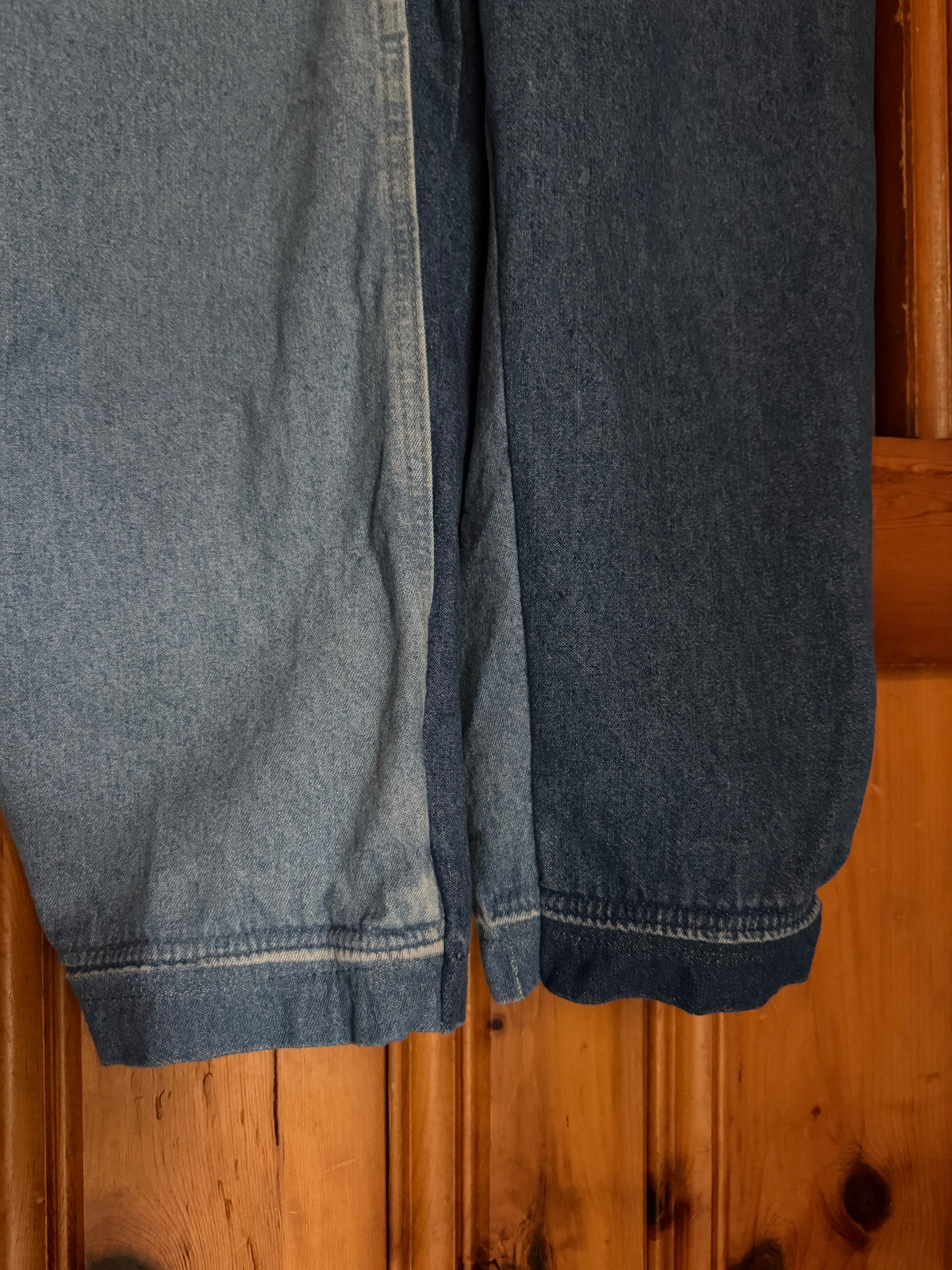 Close-up of a pair of blue denim jeans hanging against a warm wooden door, showing the hem and fabric texture in light and dark washes.