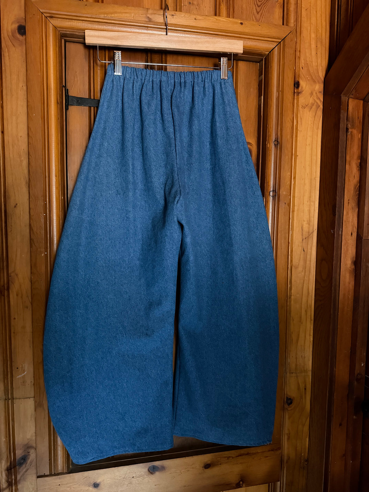 A pair of loose, wide-leg blue pants hanging from a wooden hanger on a rustic wooden door.