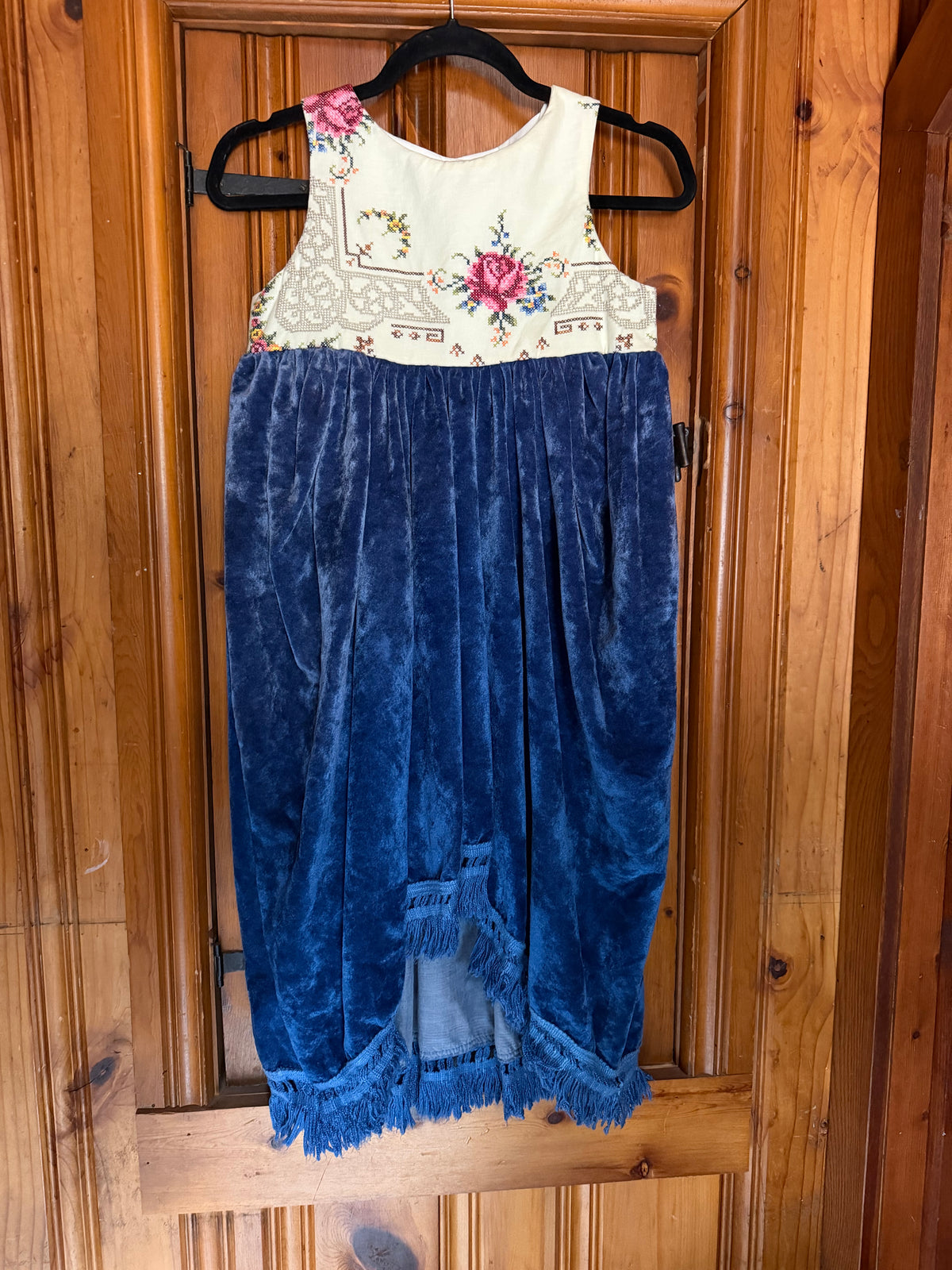A sleeveless baby dress hanging on a black hanger against a wooden door. The dress has a cream bodice with pink and blue floral embroidery and a blue velvet skirt with fringe trim.