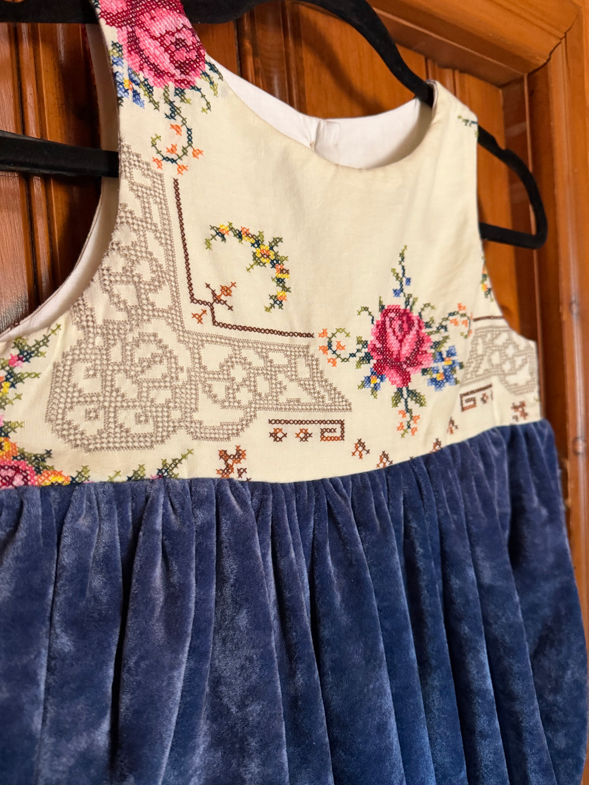 A sleeveless dress hanging on a black hanger against a wooden door. The top portion is cream with embroidered floral and geometric patterns, and the lower portion is a dark blue velvet skirt.