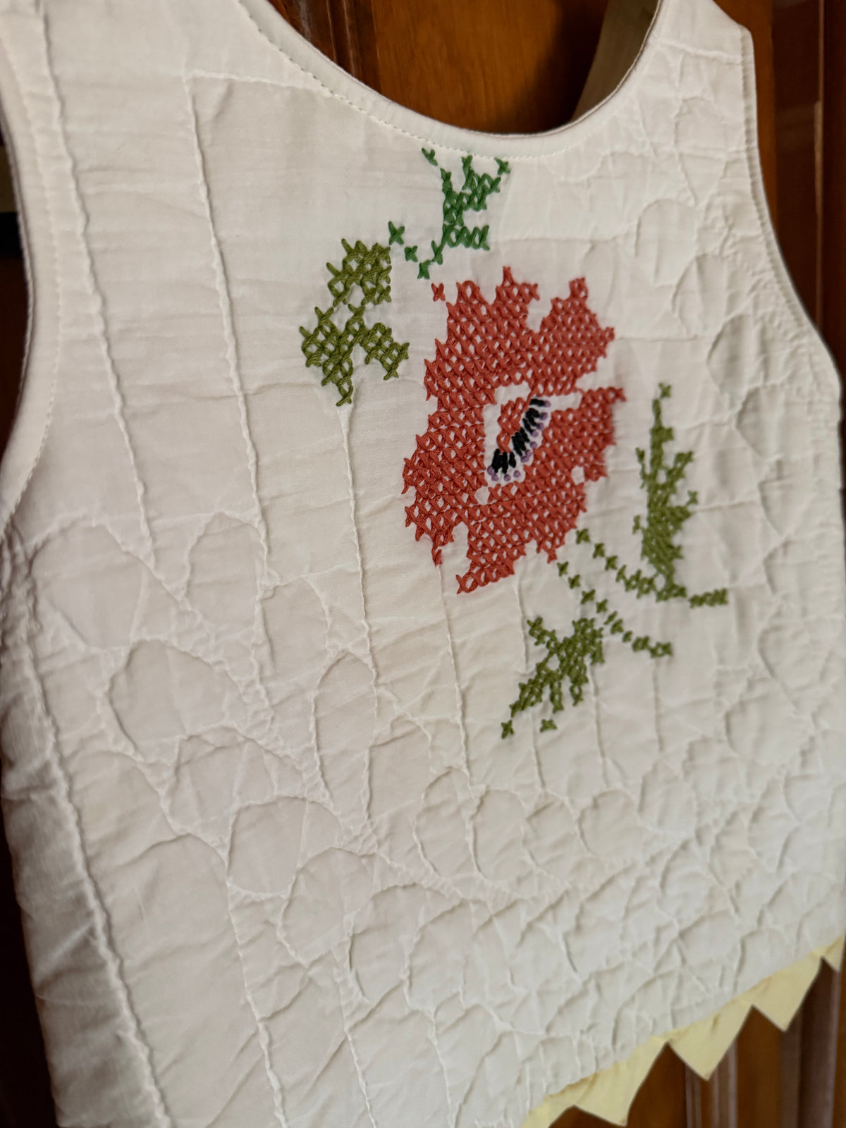 A close-up of a white quilted sleeveless garment, likely a bodice or apron panel, featuring a red and green embroidered floral motif on the chest.