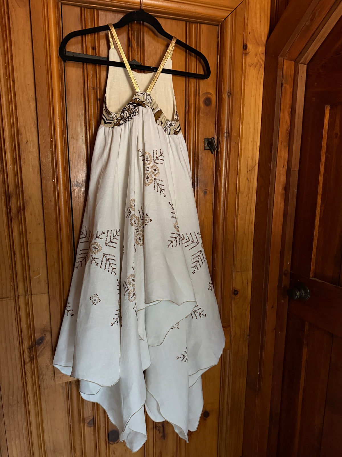 A cream-colored halter dress with brown embroidered leaf patterns and a gold-toned embellished bodice, hanging on a black hanger against a wooden door.