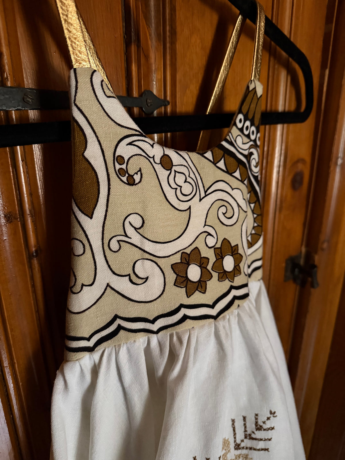 A sleeveless dress or apron-style garment hanging on a black hanger against a wooden door. The upper section has a beige base with white and brown ornamental swirls and floral motifs, while the lower section is white with subtle embroidery.
