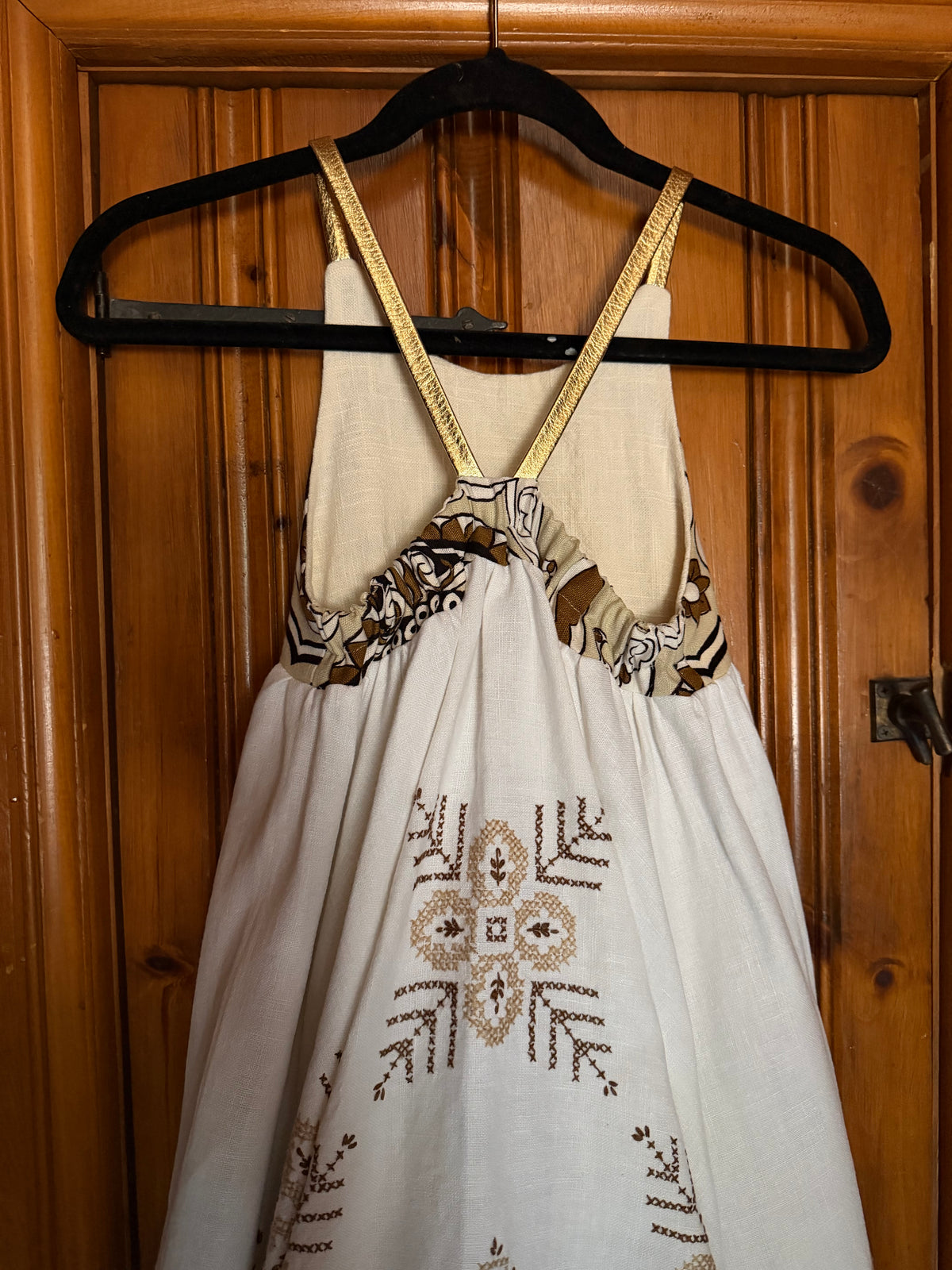 A sleeveless cream-colored dress with brown and black geometric embroidery and patterned trim, hanging on a black hanger against a wooden door.