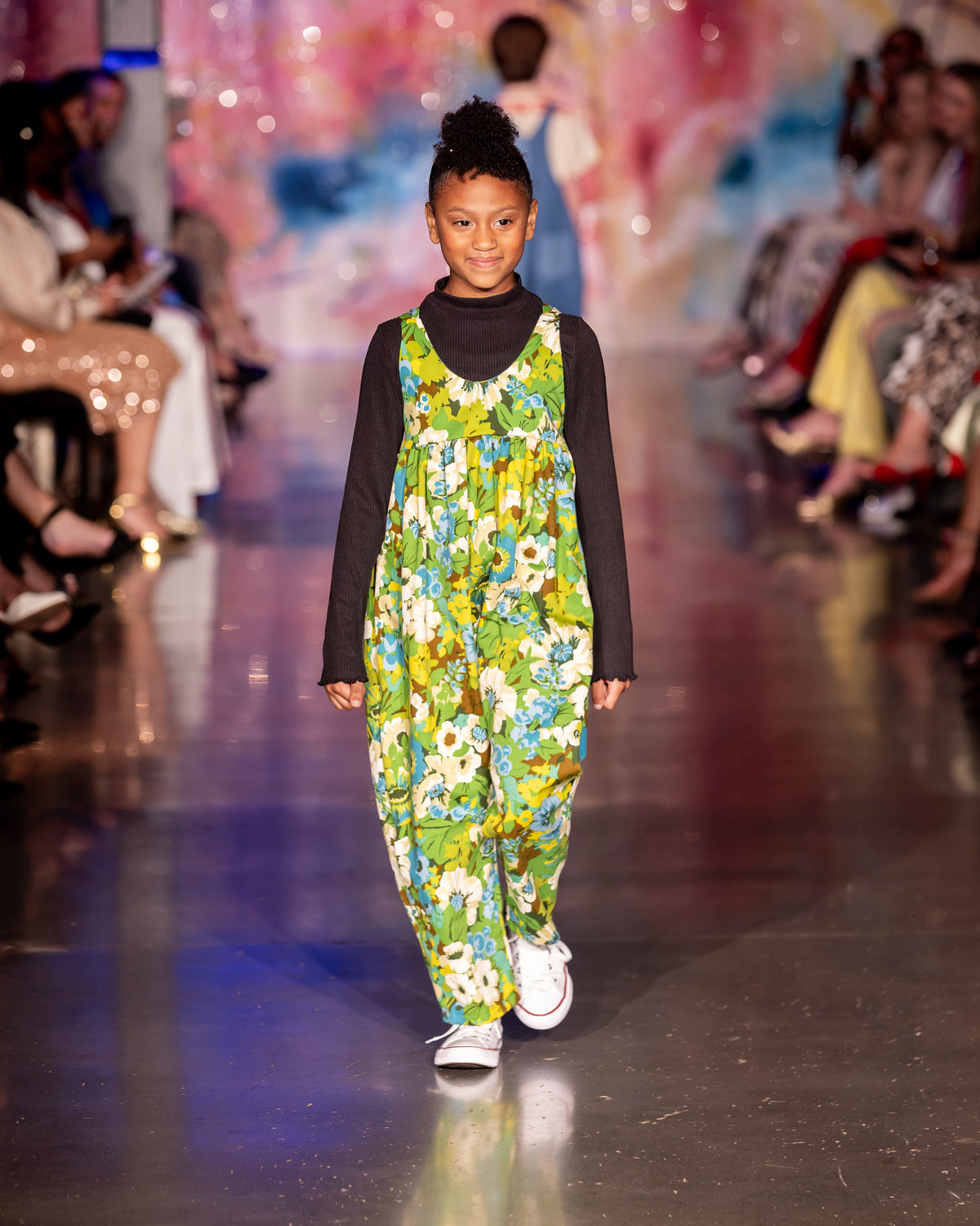 Girl walks runway in a floral green floral jumpsuit and white canvas shoes. 