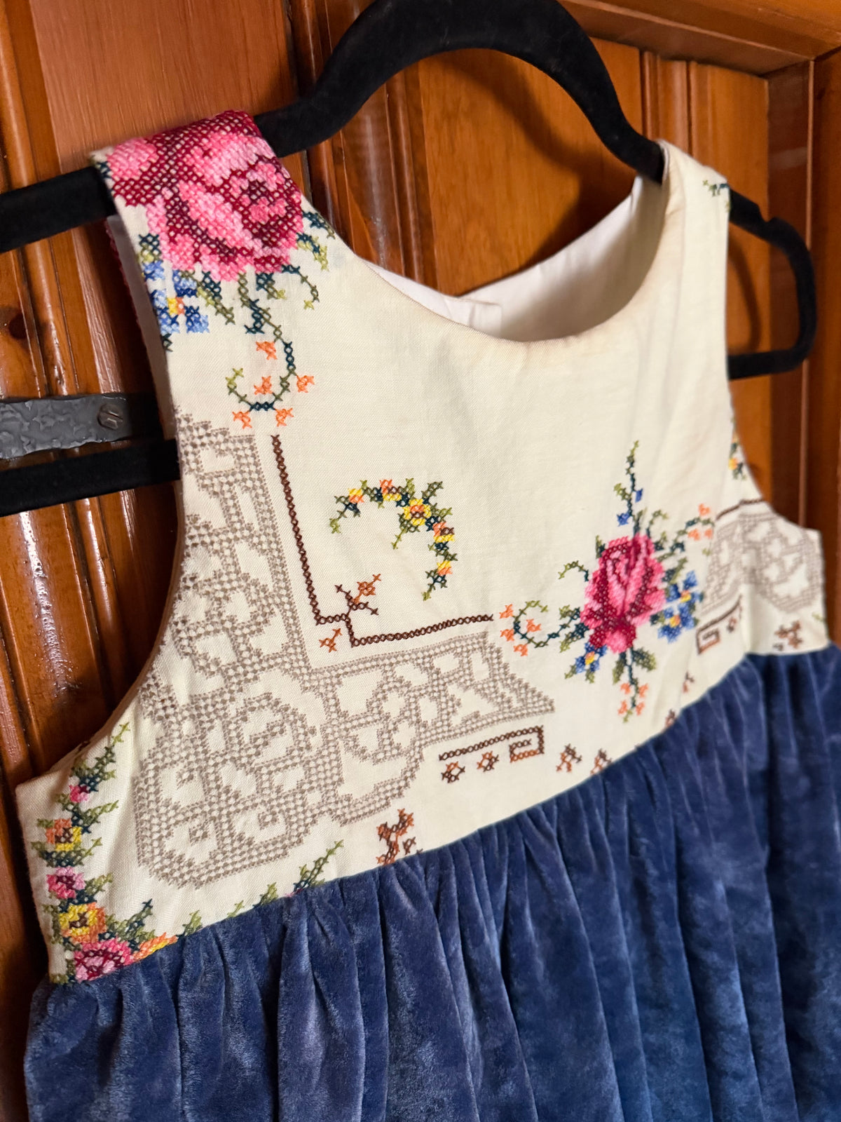A sleeveless dress or bodice with a cream embroidered upper section featuring floral and geometric cross-stitch patterns, and a dark blue gathered skirt, hanging on a black hanger against a wooden door.