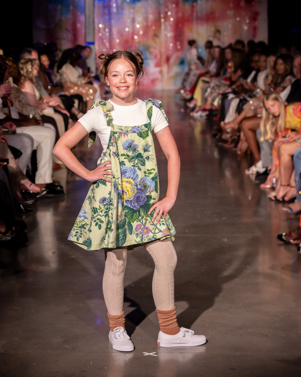 A young girl stands on a fashion runway wearing a light green floral pinafore dress over a white t-shirt, beige tights, and white sneakers. The runway is lined with seated spectators under bright stage lighting.
