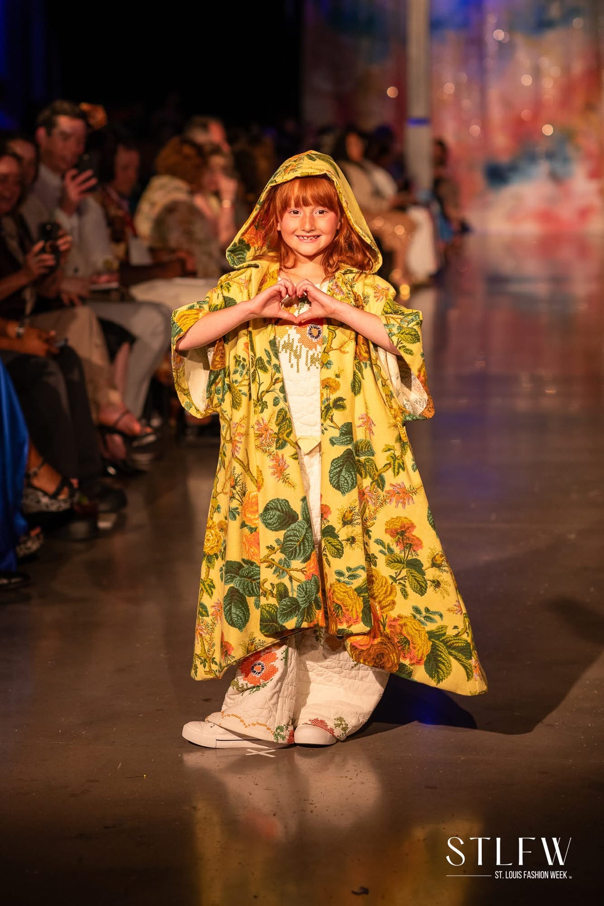 A young girl with red hair walks down a fashion runway wearing a bright yellow hooded cape with a green and orange floral print, paired with white pants and white boots. The runway is lined with an audience, and the background is dark with stage lighting.