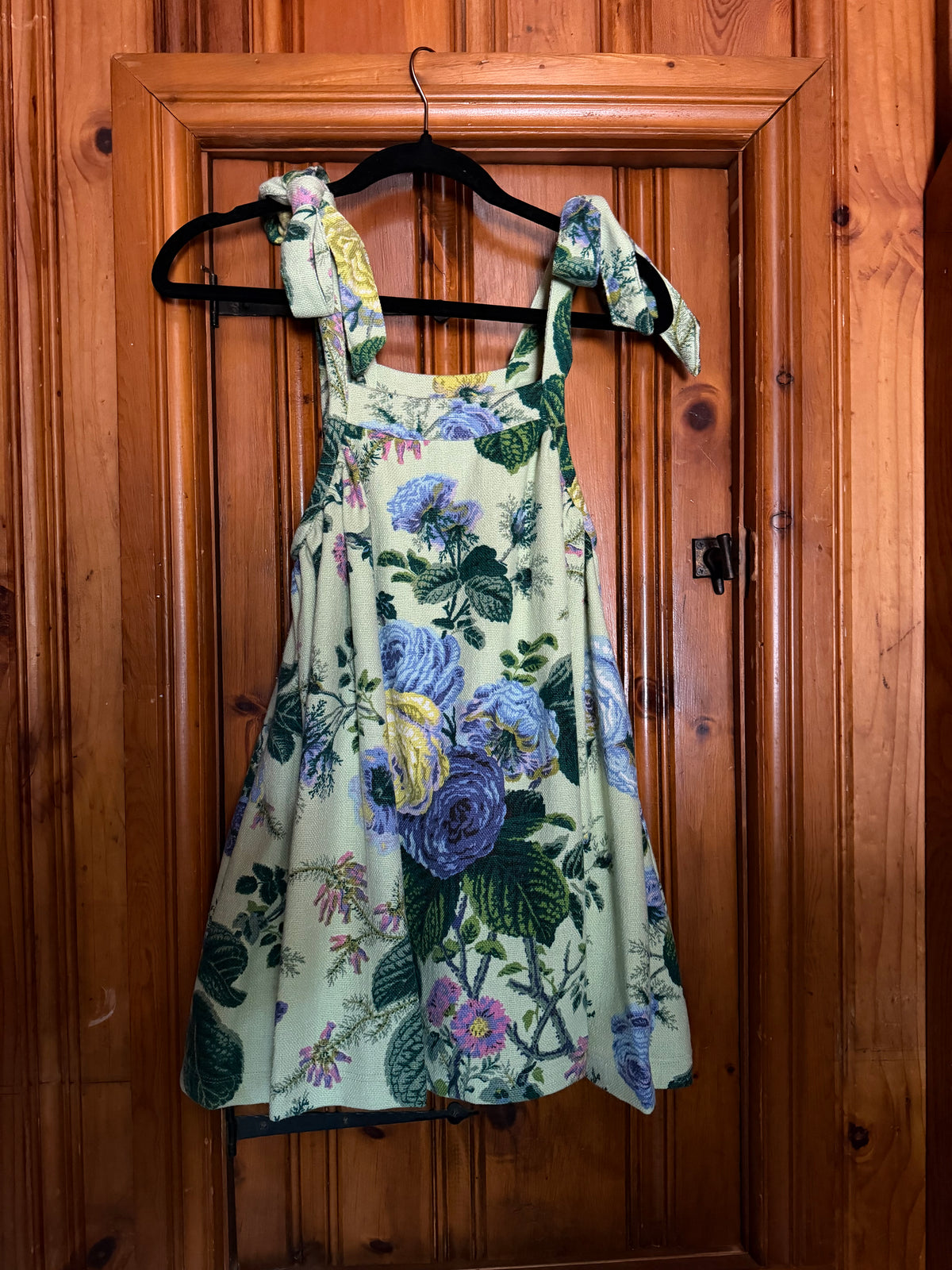 A sleeveless floral dress with tie-shoulder straps hangs on a black hanger against a wooden door. The dress has a light sage green base with a large print of blue, purple, and green flowers.