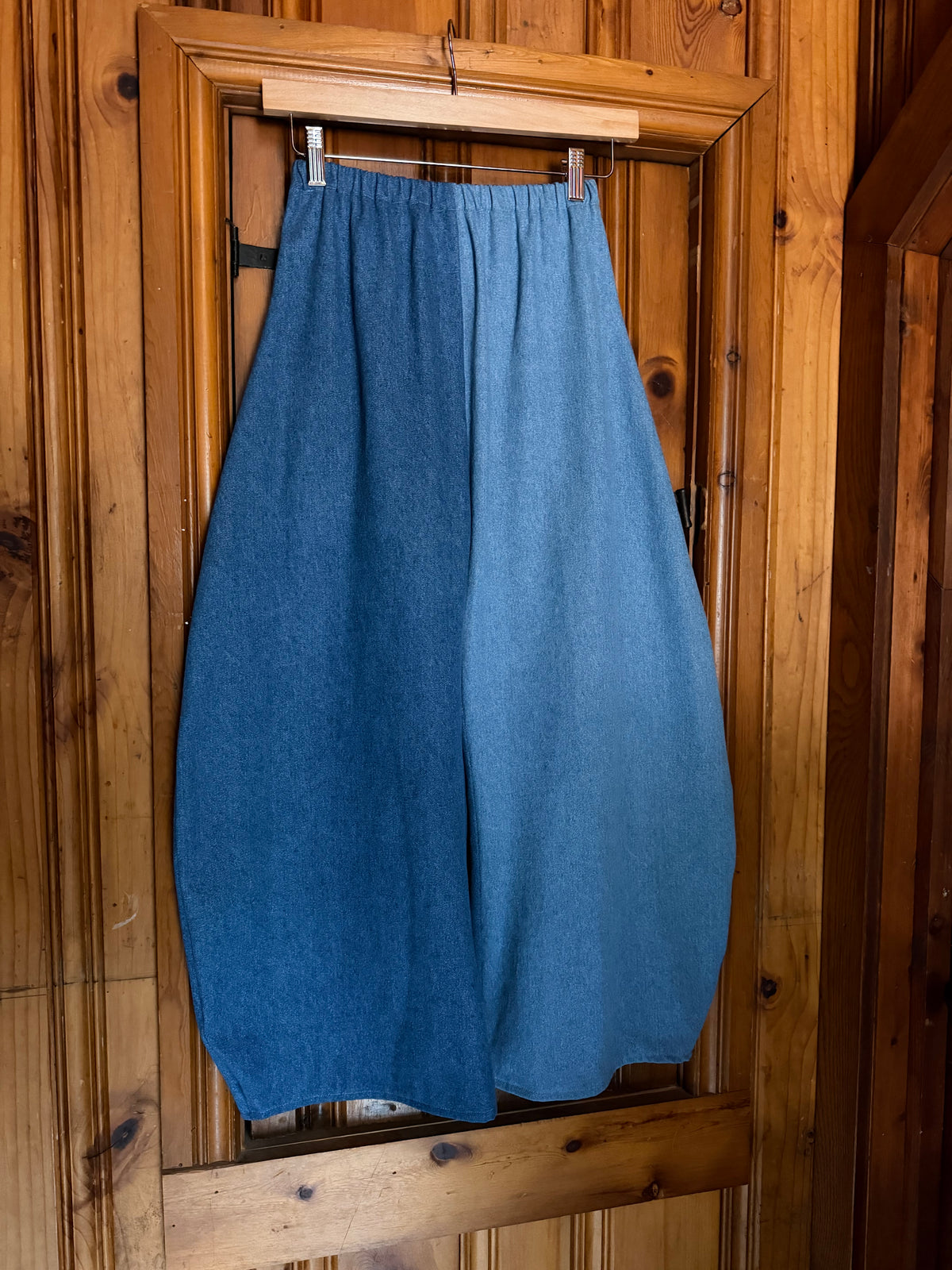 A pair of loose, balloon-style blue woolen pants or harem shorts hanging from a wooden hanger on a rustic wooden door.