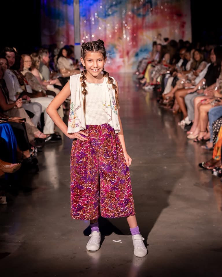 A young girl models a colorful outfit on a fashion runway, featuring a white sleeveless top with floral embroidery, a white vest, and wide-leg purple cropped pants with a vibrant pink, purple, and gold abstract pattern. The runway is lined with seated fashion industry observers under bright overhead lighting.