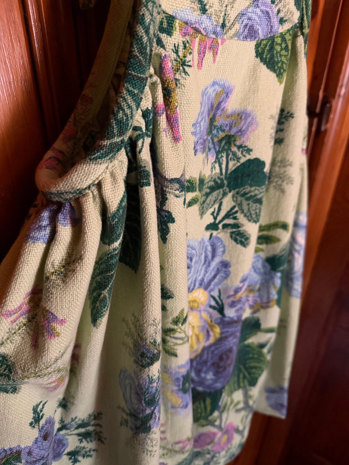 A close-up of a cream-colored woven textile, likely a tapestry or heavy fabric panel, featuring a floral pattern of purple flowers and green leaves, hanging against a dark wooden door.