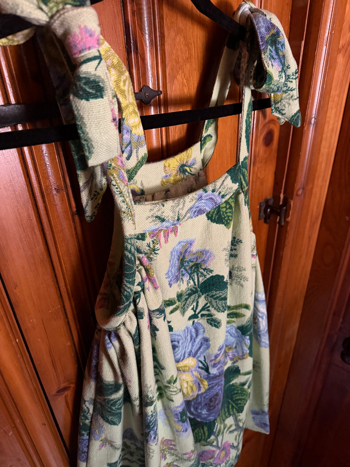 A sleeveless floral dress with tie-shoulder straps hanging on a black hanger against a warm brown wooden door background.