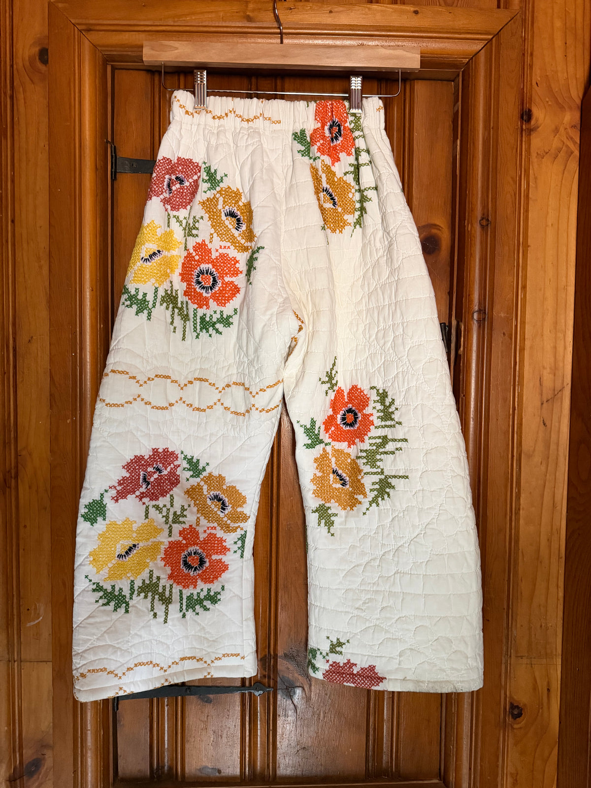 A pair of cream-colored quilted pants with colorful embroidered poppy flowers, hanging on a wooden door.