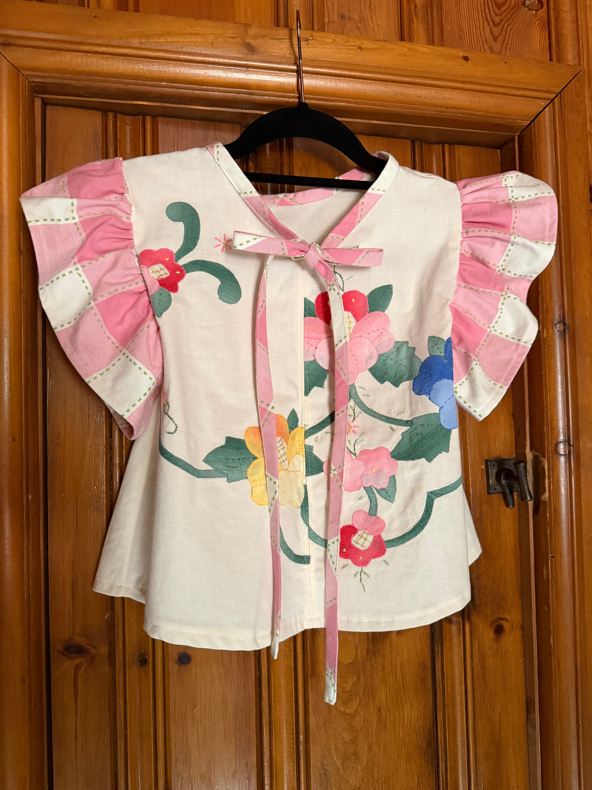 A cream-colored short-sleeve blouse with pink ruffled sleeves and a pink tie-neck bow, decorated with colorful floral embroidery in pink, red, yellow, and blue, hanging on a black hanger against a wooden door.