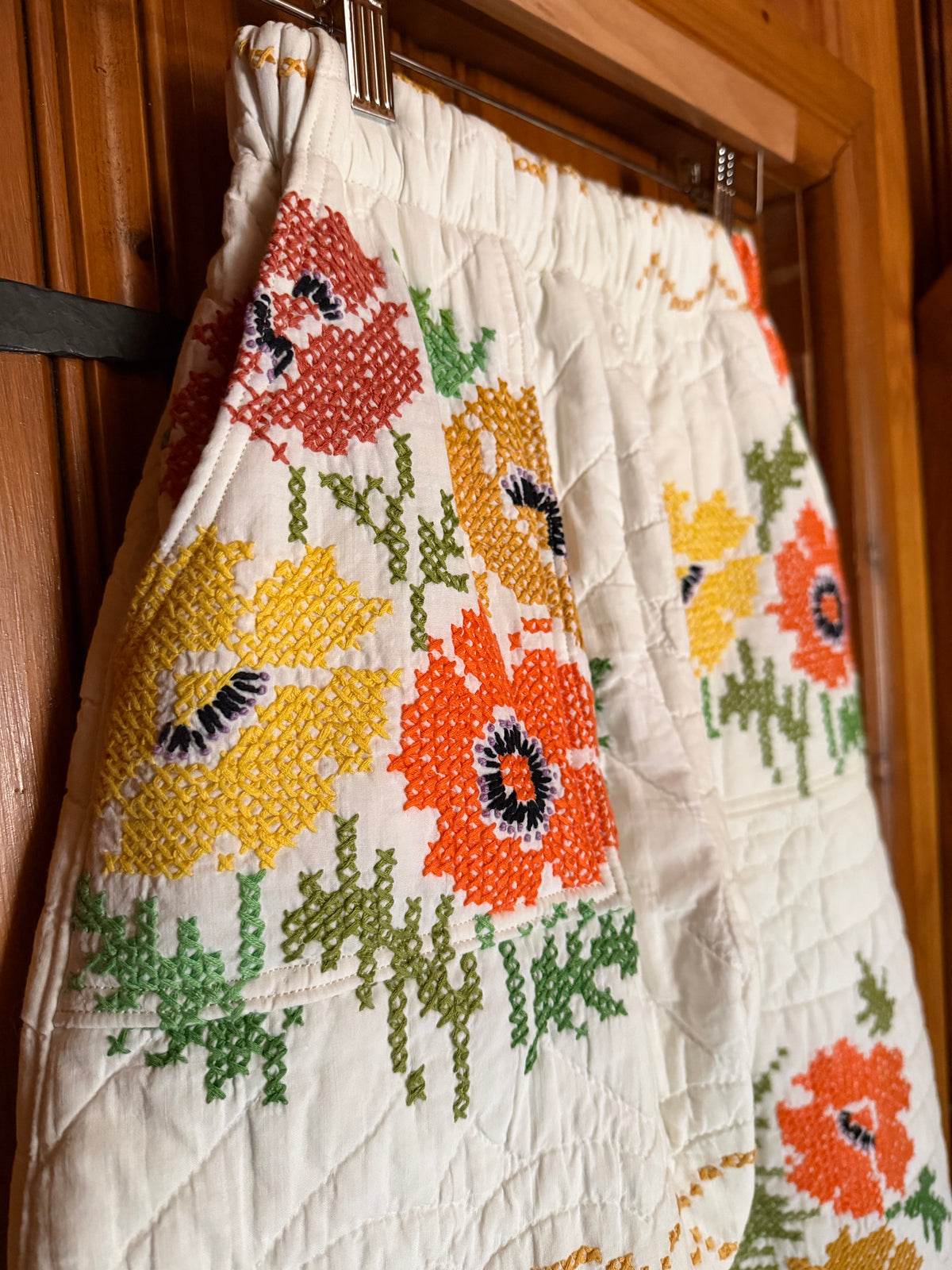A quilted white skirt with a colorful embroidered poppy floral pattern, hanging on a wooden hanger against a wooden door background.