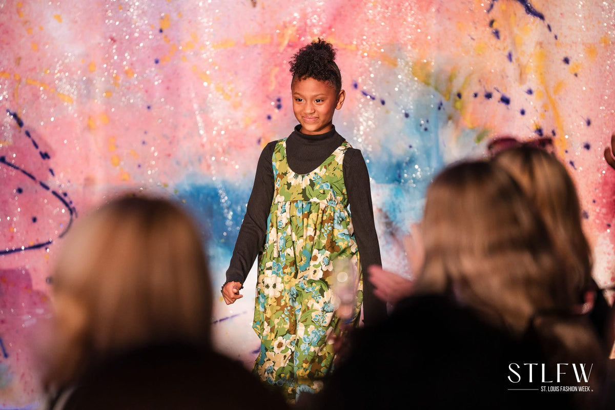 Girl poses at start of fashion runway in a floral green floral jumpsuit in front of pink and blue graffiti canvas backdrop. 