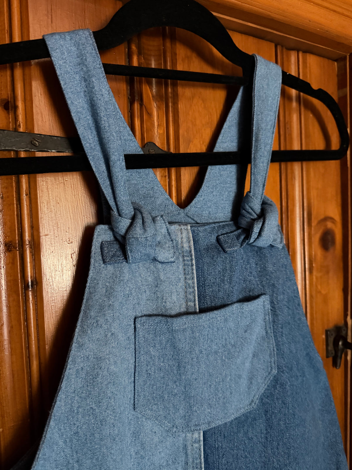 A light blue denim overall hanging on a black hanger against a warm wooden door background.