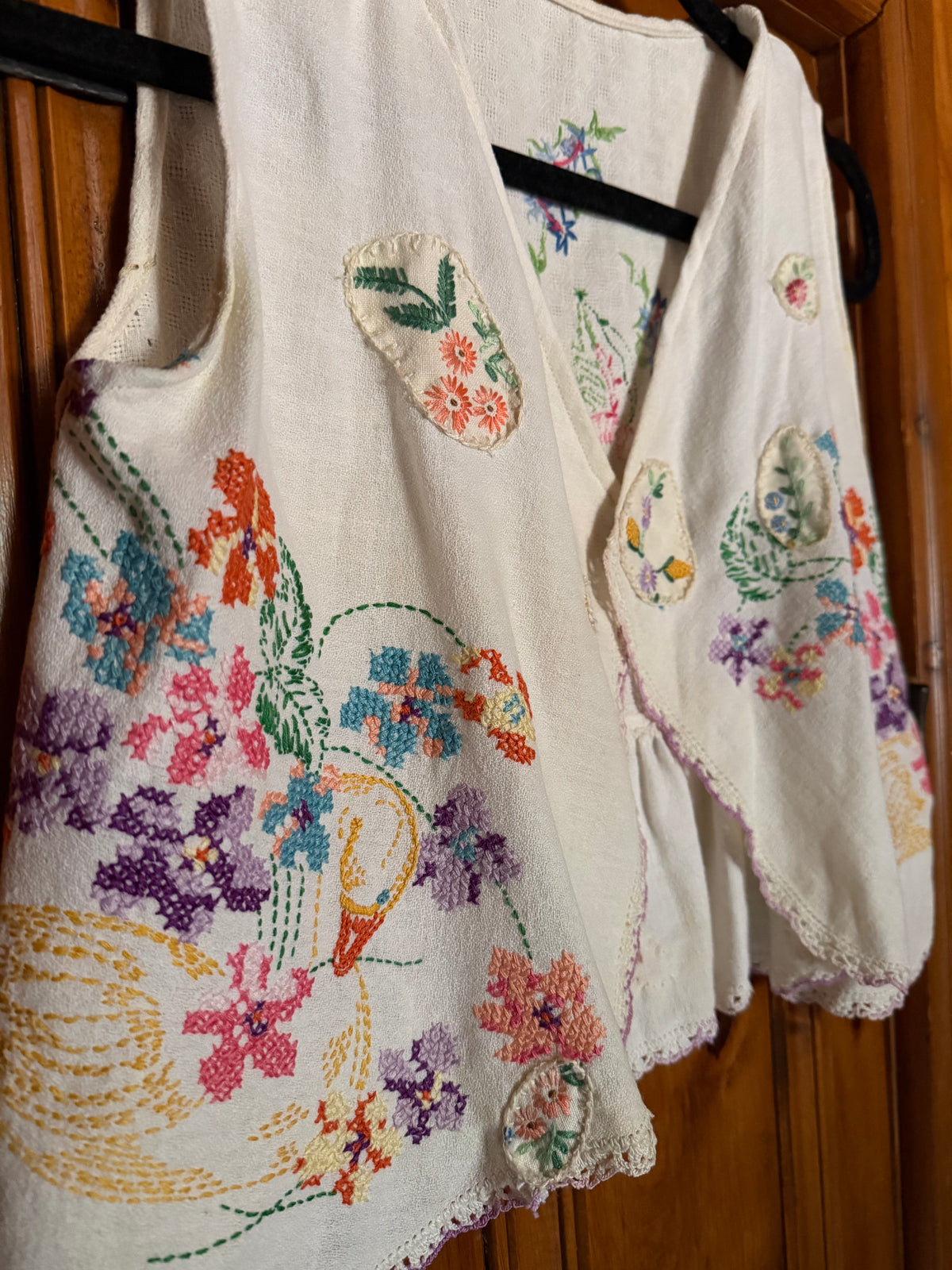A cream-colored linen vest decorated with colorful hand-embroidered floral motifs and a large bird design, hanging on a black hanger against a wooden door background.