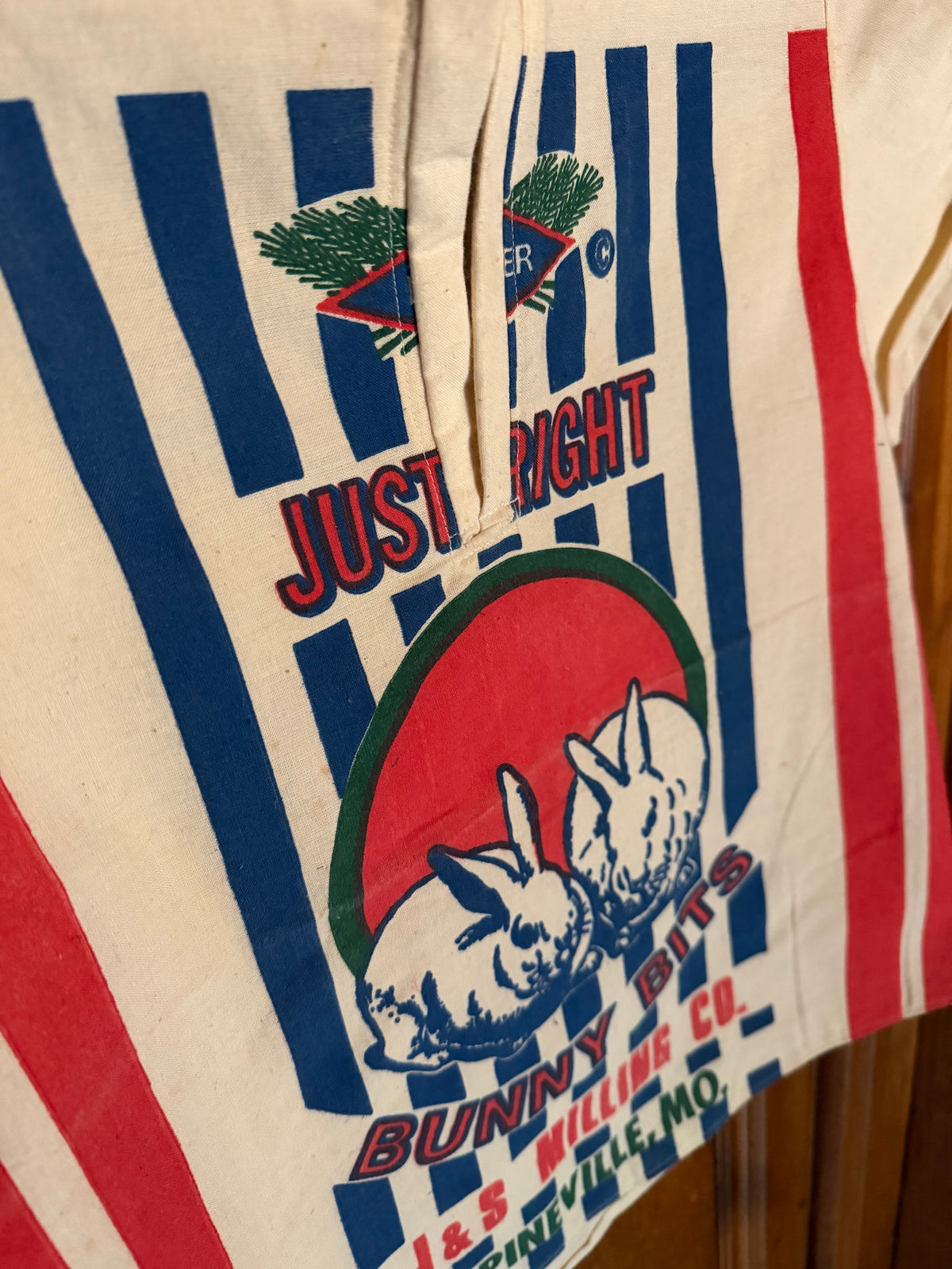A vintage cream-colored cotton shirt or apron with bold blue and red vertical stripes, featuring a large graphic of two white rabbits inside a red circle with the text 'JUST RIGHT BUNNY BITS' and '189 MILLION CO. ENSWILLLIE, MO.