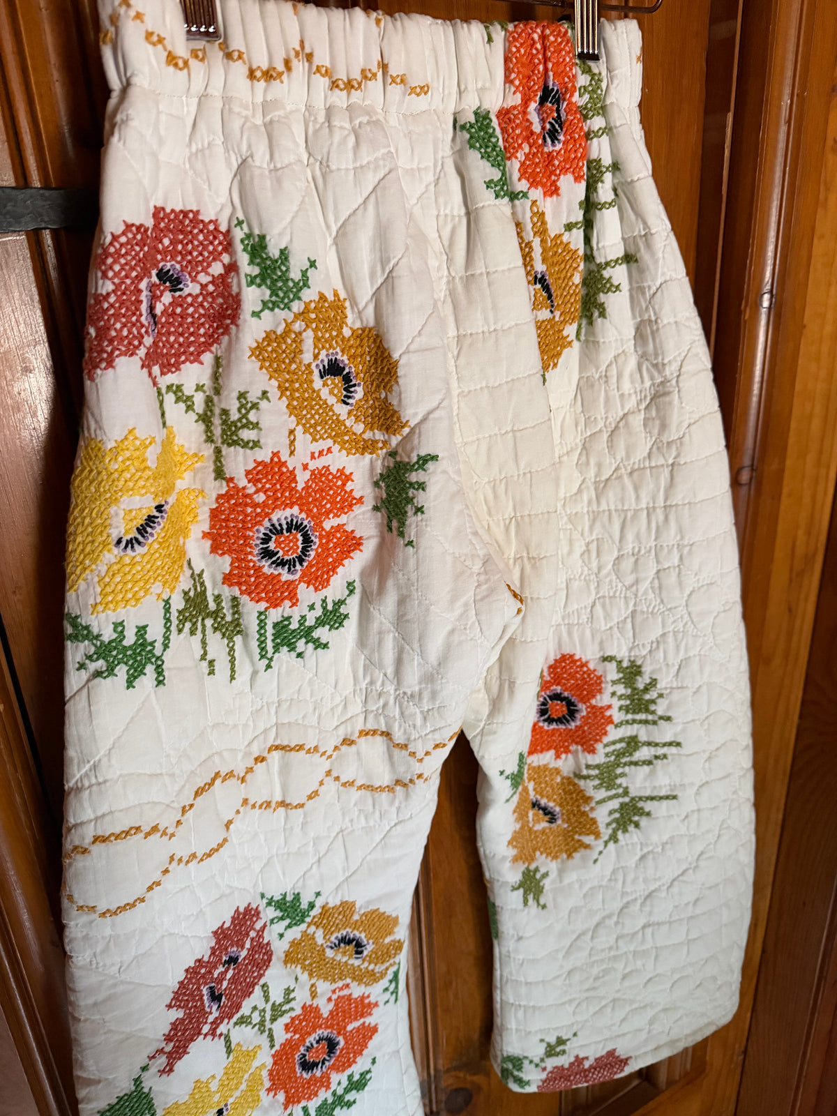 Cream-colored quilted trousers with a colorful embroidered floral pattern, hanging on a wooden door.