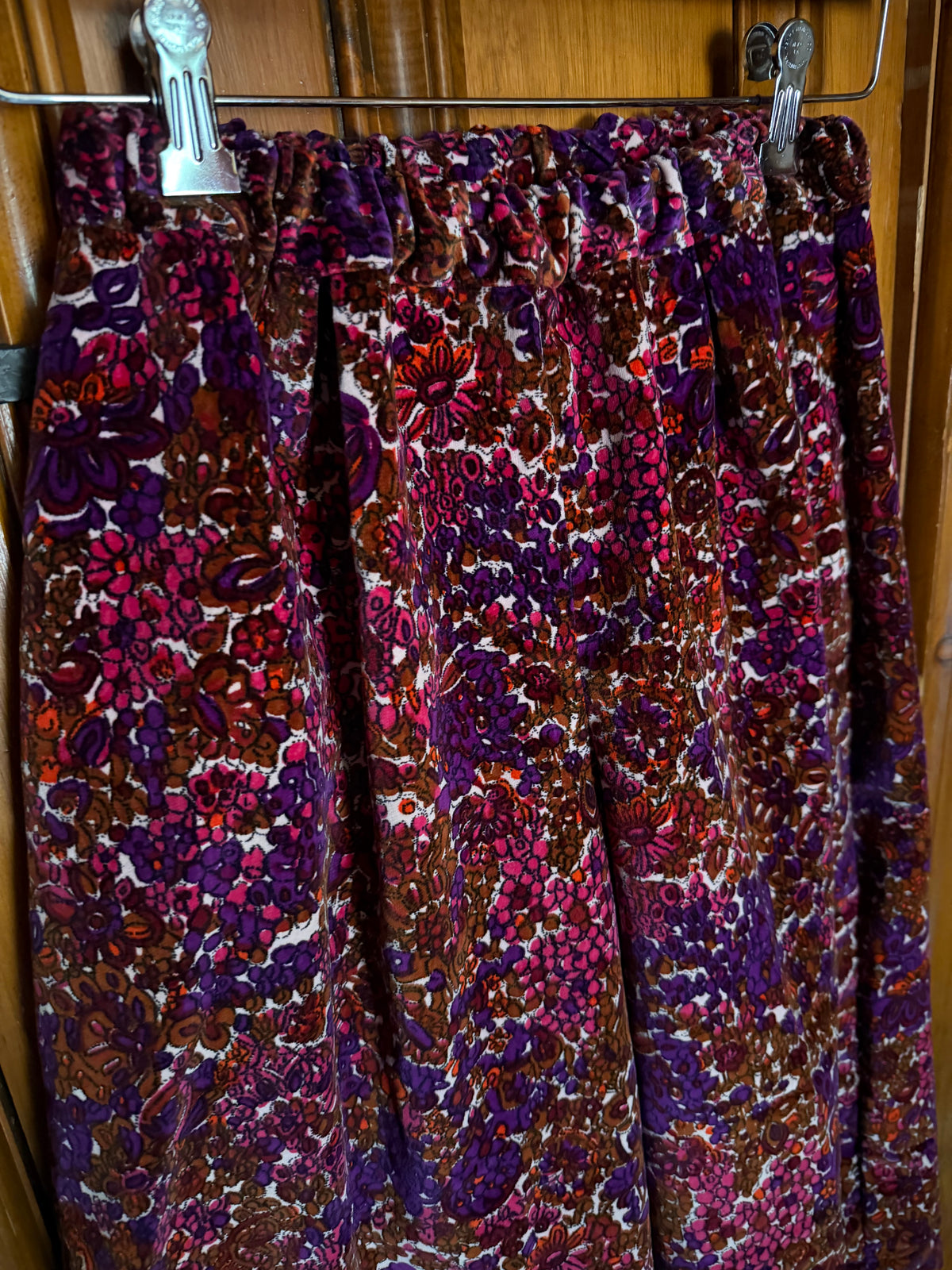 A pair of gaucho wide-leg trousers with an intricate, dense floral and paisley pattern in purple, magenta, brown, and white, hanging from metal clips on a wooden door.