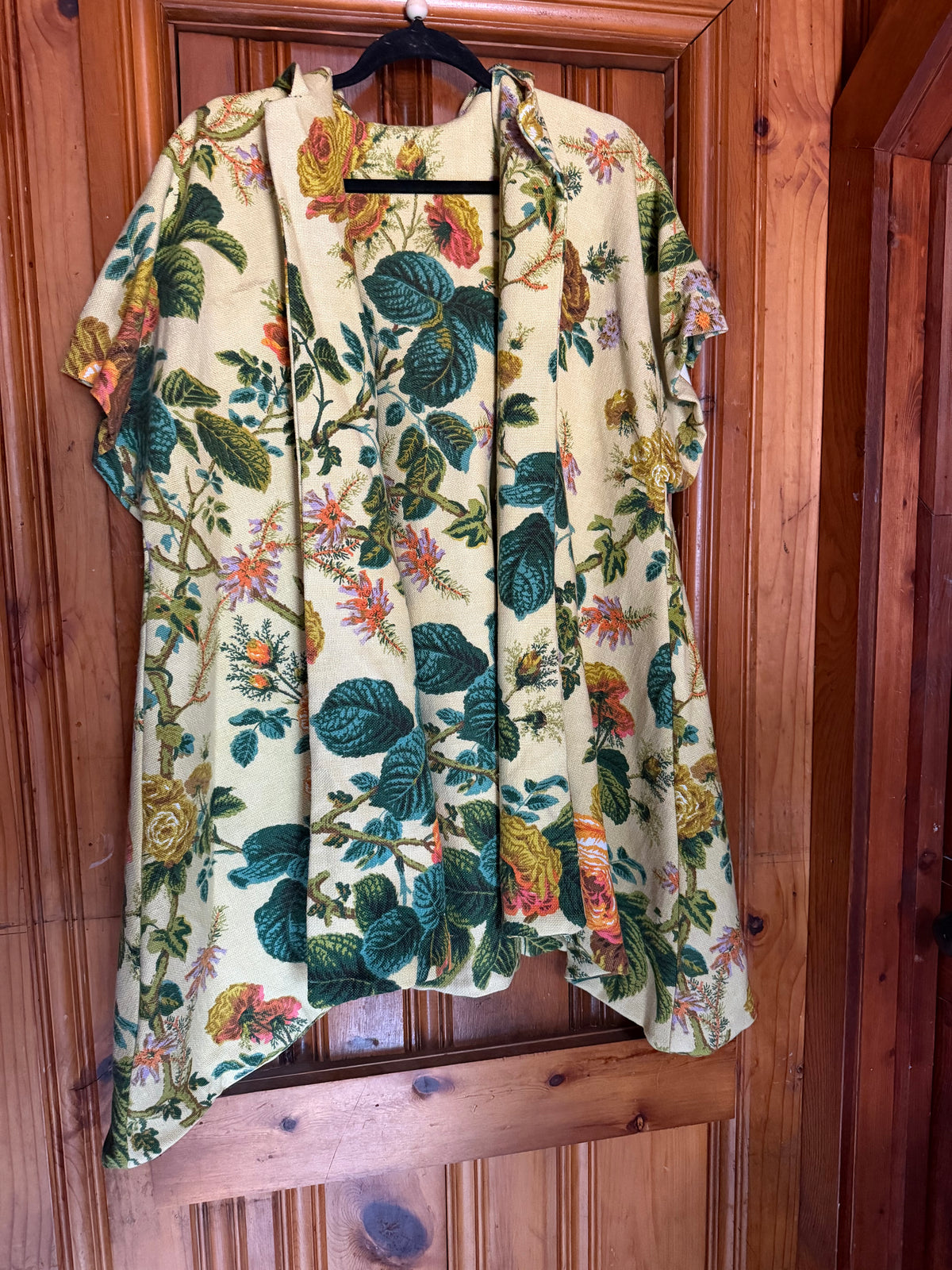 A short-sleeve open-front kimono-style top with a vintage botanical print, hanging on a black hanger against a wooden door.
