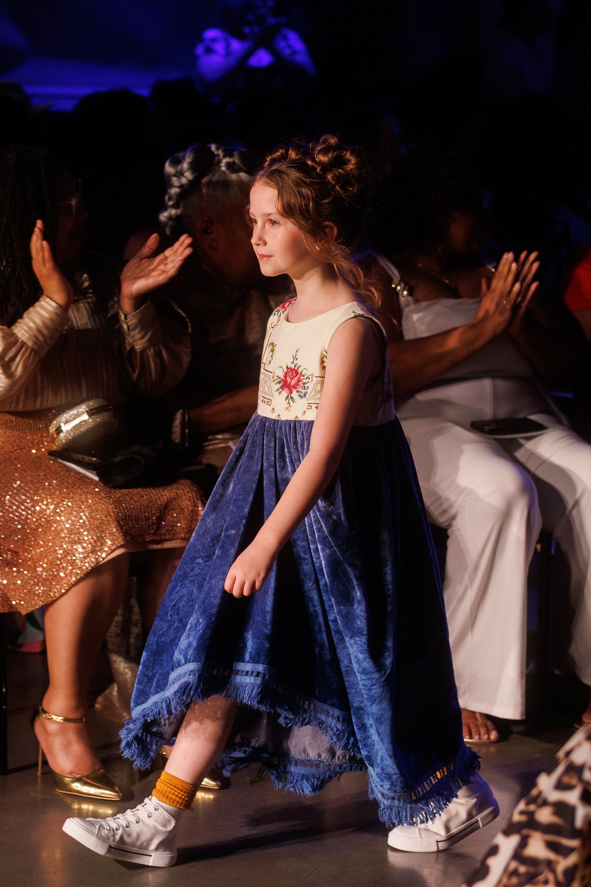 A young girl walks down a fashion runway wearing a sleeveless dress with a cream bodice featuring a red floral embroidery and a blue velvet skirt with fringe. She is walking on a dark runway with audience members applauding in the background.