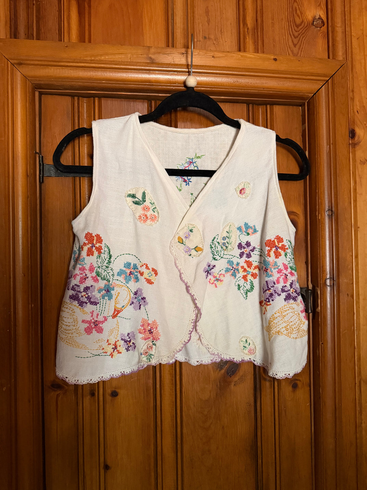 A sleeveless cream-colored linen vest with colorful floral embroidery, hanging on a black hanger against a wooden door.