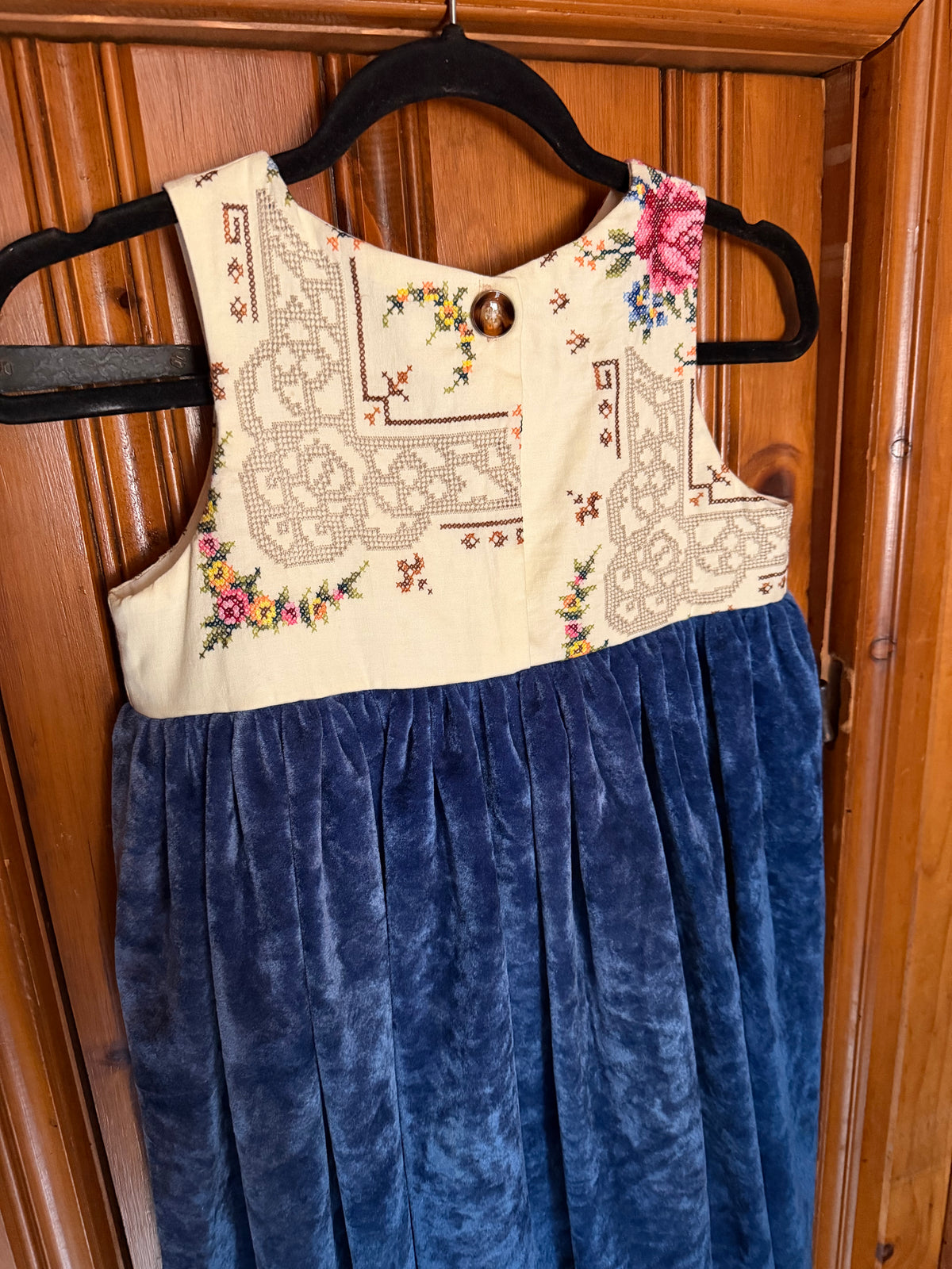 A sleeveless vintage-style dress hanging on a black hanger against a wooden door. The bodice is cream with embroidered floral and geometric patterns and a small brown button at the neckline.