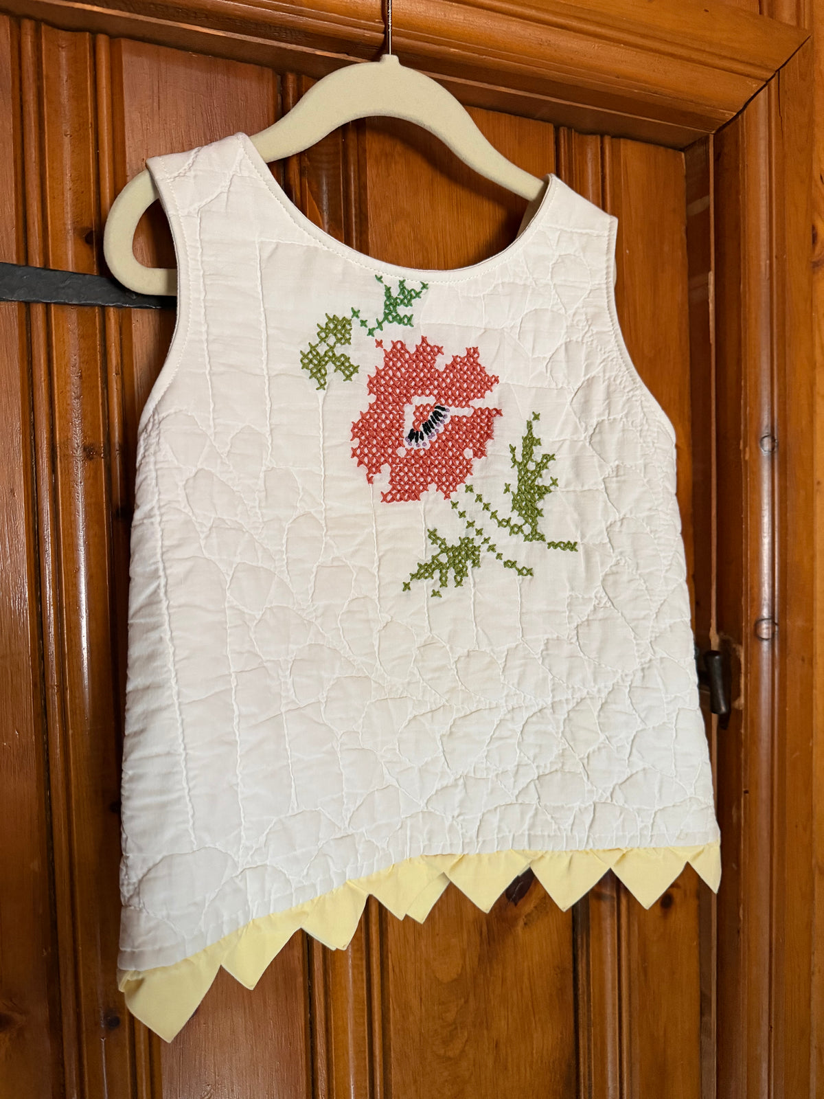 A sleeveless white quilted top with a red and green floral embroidery on the chest and a yellow scalloped hem, hanging on a wooden hanger against a wooden door.