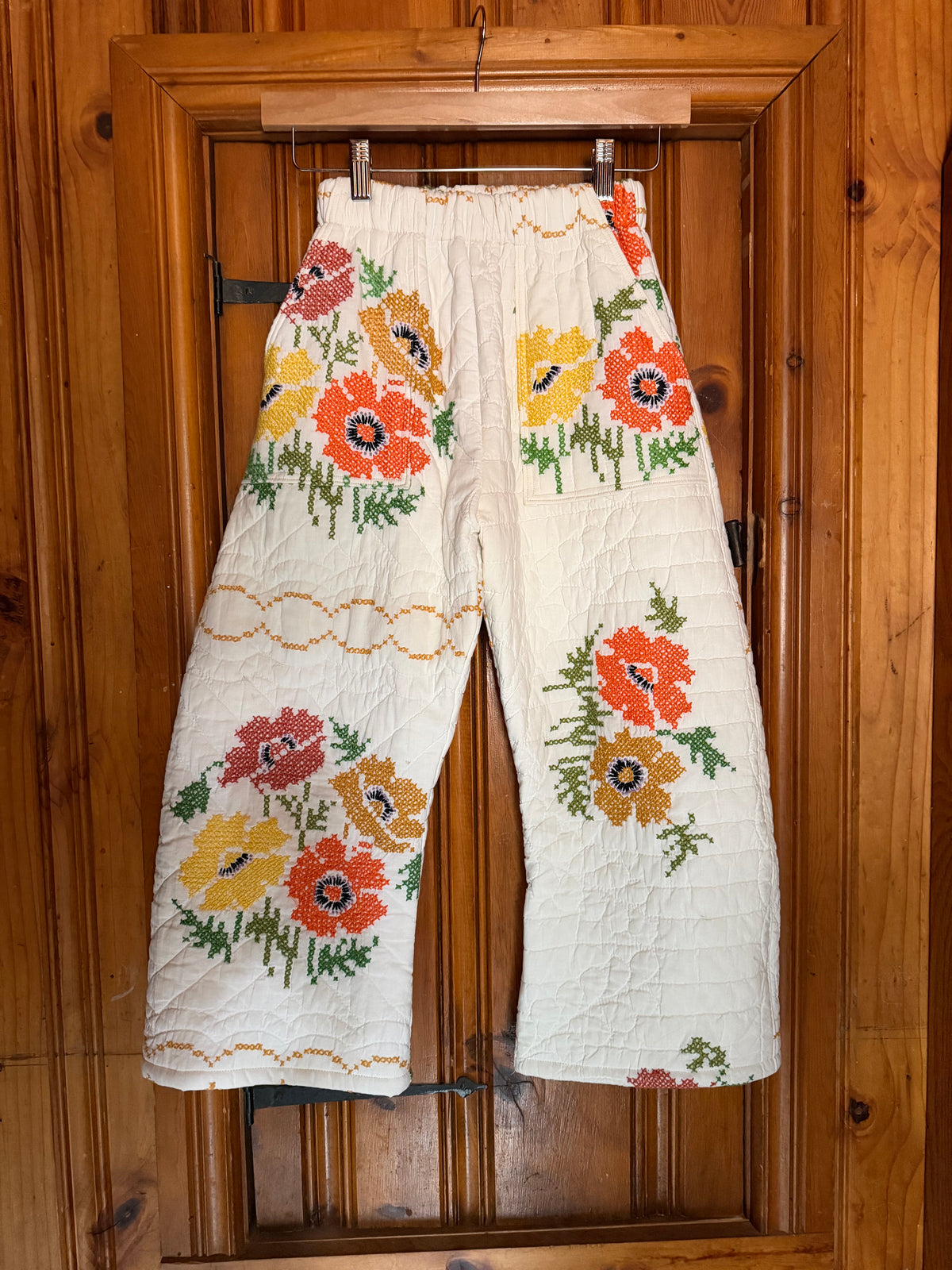 A pair of off-white cropped pants with colorful embroidered poppy flowers in orange, yellow, and red, hanging from a wooden hanger against a wooden door.