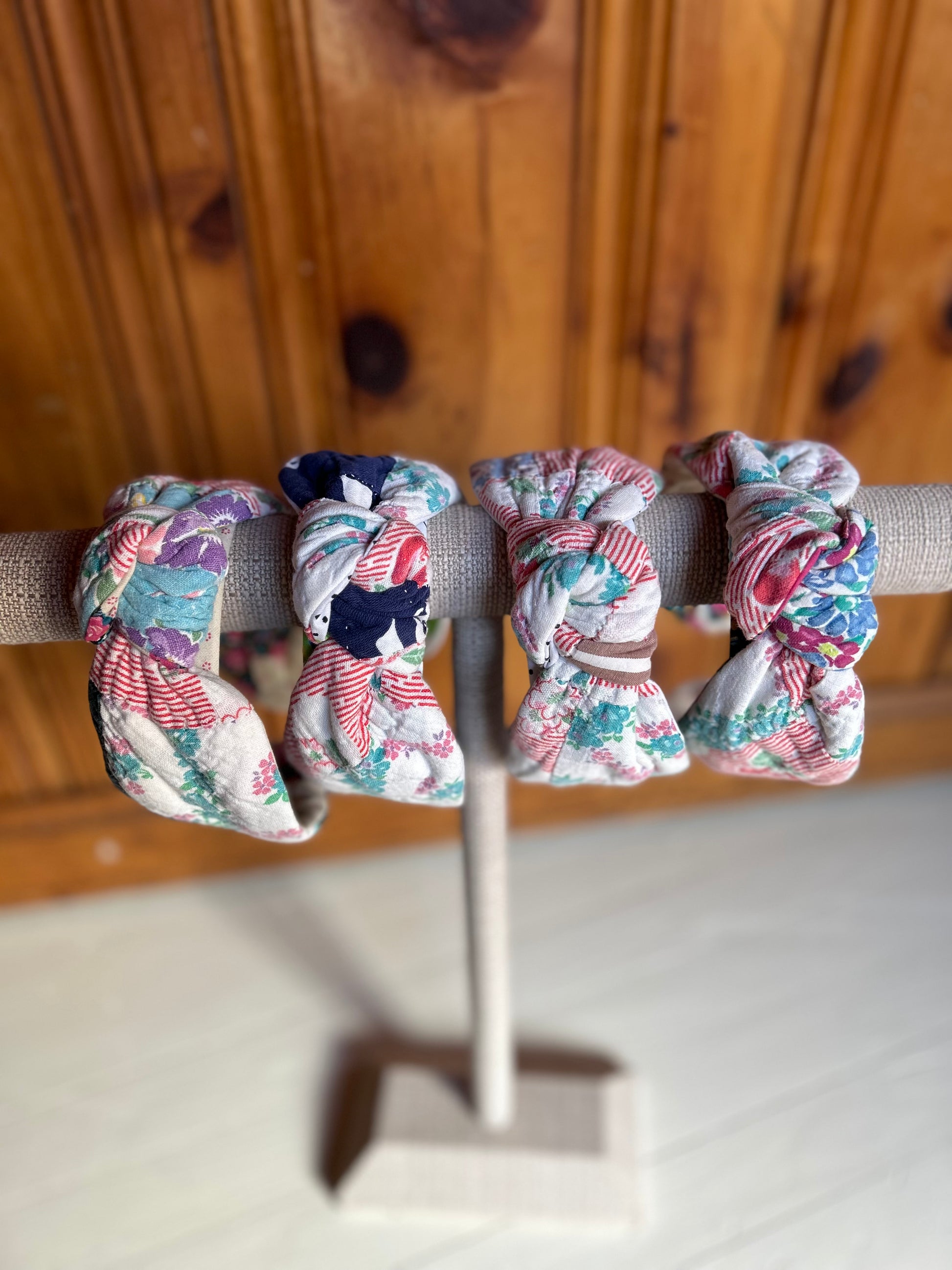 Image is of four knot headbands constructed with a vintage multi color quilt. Primary colors consist of a base color of white with red, greens and blues. The headbands are hanging on a burlap covered necklace holder.