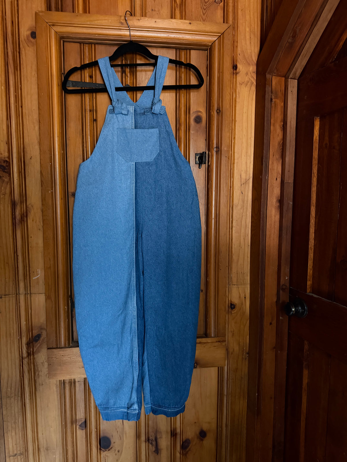 A pair of blue denim bib overalls hanging on a black hanger against a rustic wooden door.