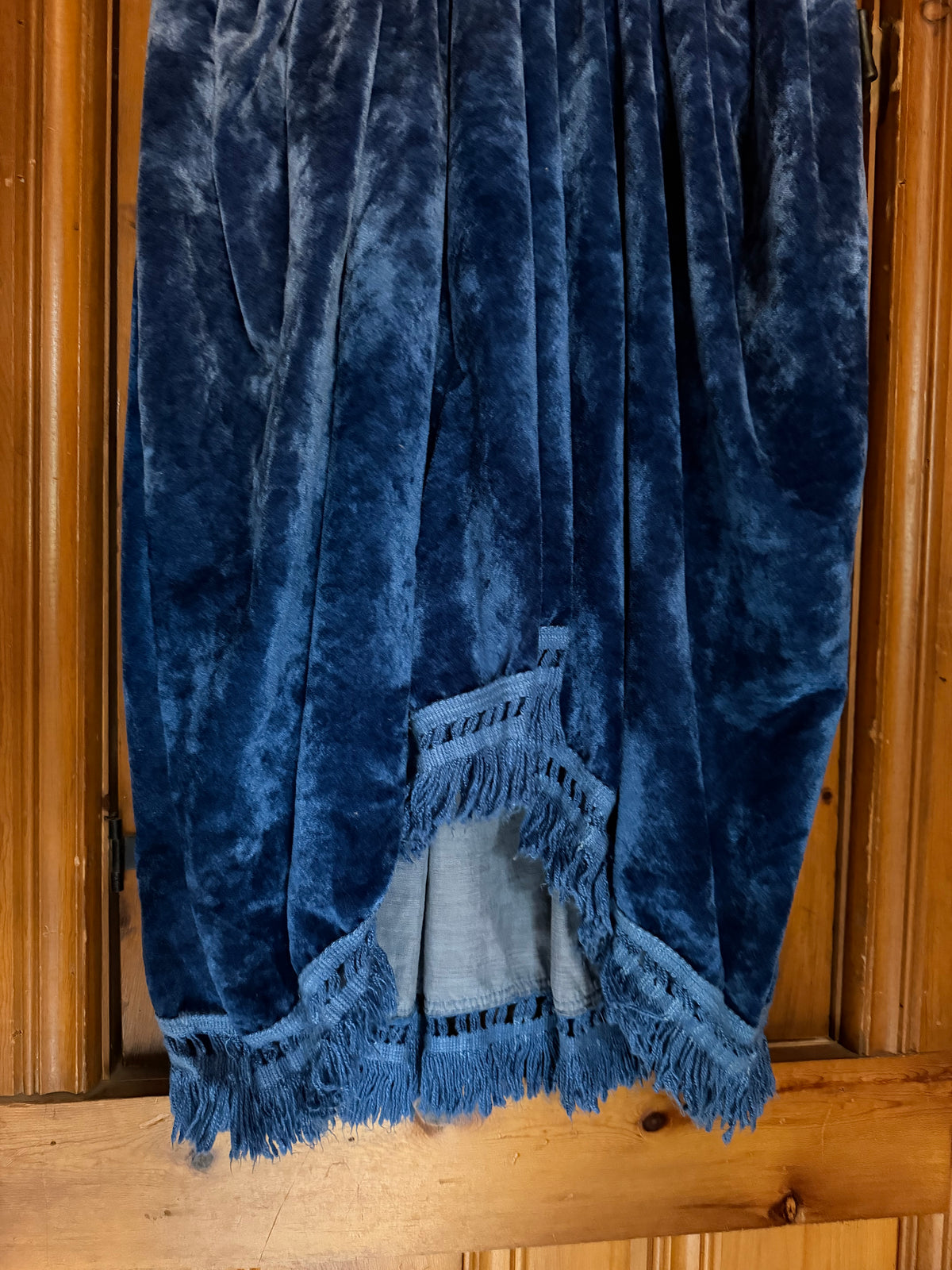 A dark blue velvet fabric skirt draped over a wooden surface, featuring a fringed hem and a lighter blue inner lining visible at the opening.